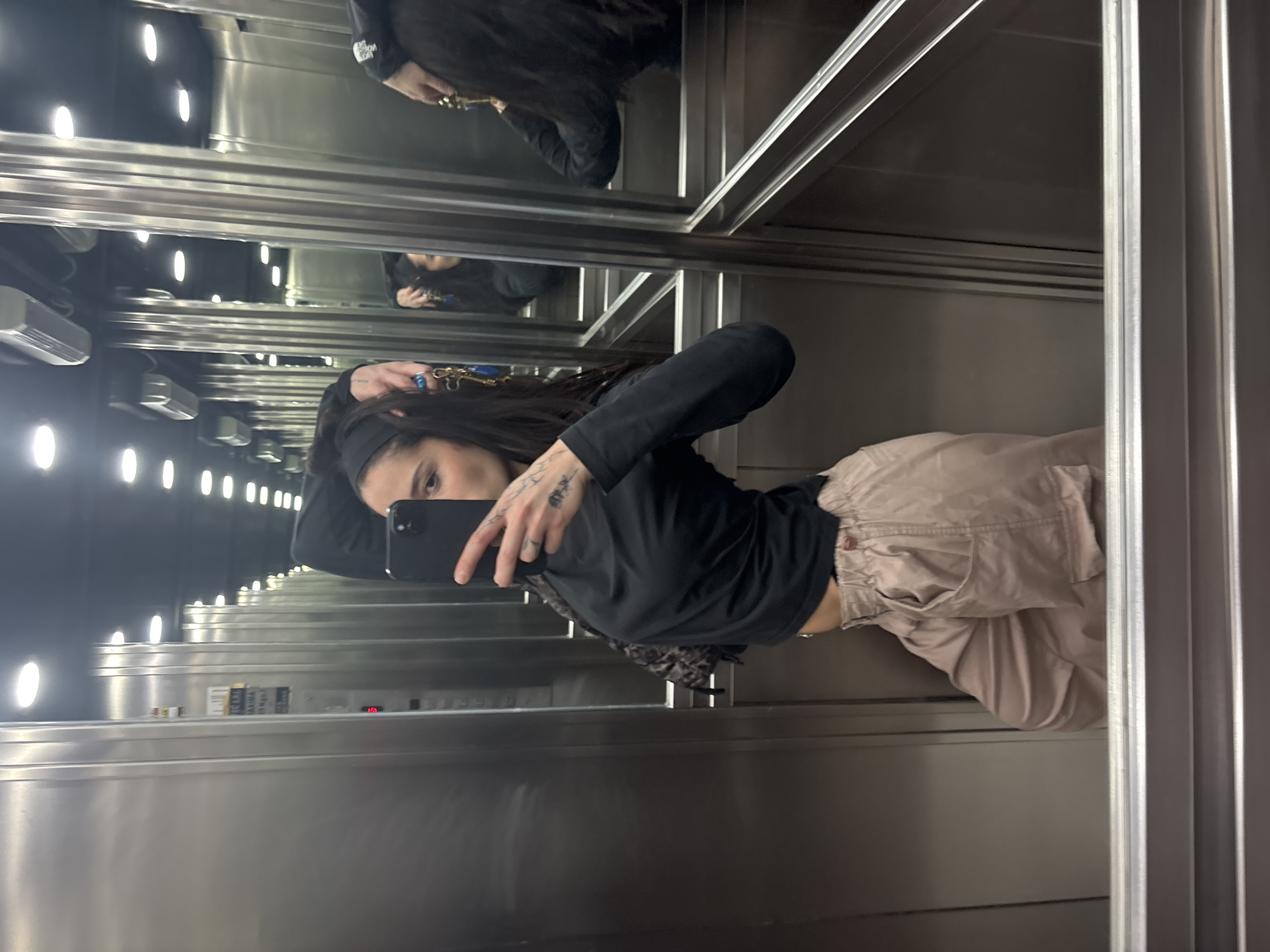 The image, rotated 90 degrees clockwise, captures a young woman taking a mirror selfie inside a modern elevator. The woman, with long dark hair partially pulled back by a dark headband, is wearing a long-sleeved black top and light brown or khaki cargo pants. She holds a black smartphone, likely an iPhone, in her left hand, extending it to capture her reflection in the elevator's mirrored walls. Visible on her left hand and forearm are several tattoos, and she wears a prominent ring on her right hand's middle finger. A patterned, possibly leopard print, fabric is visible peeking out from under her left arm.
The elevator interior is characterized by sleek, brushed metallic walls and ceiling, providing highly reflective surfaces that multiply the image of the woman and the elevator's internal lights. Bright, oval-shaped recessed lights illuminate the space. In the background, part of the elevator's control panel is visible. A digital display shows the number "10", indicating the current floor. Above it, a sign with small text is partially readable, showing "MAX Kg" and a number (possibly 1350) along with "MAX Persons" and a number (possibly 18), indicating the elevator's capacity. The setting is consistent with a contemporary urban building in Buenos Aires, Argentina. The woman's expression is neutral as she looks directly into the phone's camera.