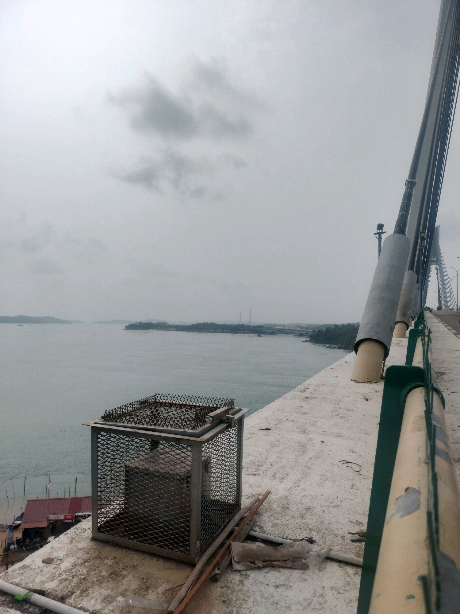 The image depicts a view from a bridge, likely part of the Trans-Barelang Bridge system in Batam, Indonesia, given the distinctive cable-stayed design and the coastal landscape with islands. The foreground features a concrete walkway of a bridge, with a metal cage-like structure, possibly a ventilation or equipment enclosure, to the left. The cage is made of diamond-shaped mesh and has a wire mesh lid. Various construction or maintenance materials, including pipes and wooden planks, are scattered on the bridge surface.
A body of water stretches out into the distance, with islands and a hazy coastline visible. The sky is overcast and gray, suggesting a cloudy or rainy day. In the far distance, a suspension bridge with multiple towers can be seen, further reinforcing the identification of the location as a significant bridge in Batam. A small settlement with red-roofed buildings is situated along the water's edge on the left side of the image, with several poles extending from the water. The overall impression is one of a functional, industrial-style bridge structure in a tropical coastal environment. There are no people visible in the image.