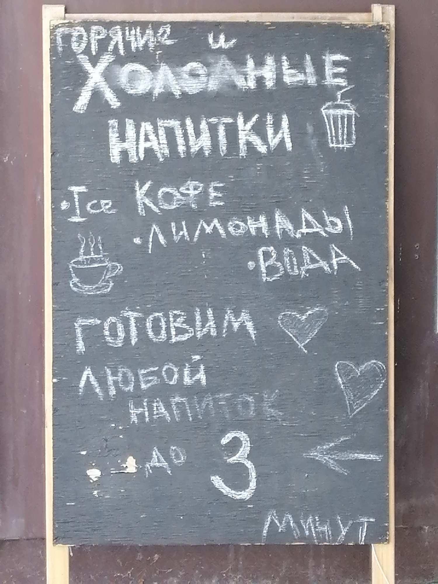 A chalk-drawn menu is displayed on a dark grey chalkboard with a light wooden frame, leaning against a brown textured surface. The chalkboard is positioned outdoors, suggesting a cafe or food stall setting.
The menu items are written in white chalk and include:
* **ГОРЯЧИЕ И ХОЛОДНЫЕ НАПИТКИ** (Hot and Cold Drinks), with an illustration of a cup with a straw.
* **• Ice Кофе** (Ice Coffee)
* **• ЛИМОНАДЫ** (Lemonades)
* **• ВОДА** (Water)
Below this, there is a drawing of a steaming coffee cup.
Further down, the menu reads:
* **ГОТОВИМ** (We Prepare)
* **ЛЮБОЙ НАПИТОК** (Any Drink)
Accompanying these are two chalk-drawn hearts.
At the bottom, there is a number:
* **3**
An arrow points to the left from the number.
* **МИНУТ** (Minutes)
The overall impression is of a casual, hand-crafted outdoor menu. The chalk lettering suggests a temporary or frequently updated offering. The presence of specific drink categories like "Ice Coffee" and "Lemonades" indicates a beverage-focused establishment. The "We Prepare Any Drink in 3 Minutes" highlights a service promise. The context of Rostov-on-Don, Russia, provides a geographical location for this scene, implying a typical urban environment within that city.