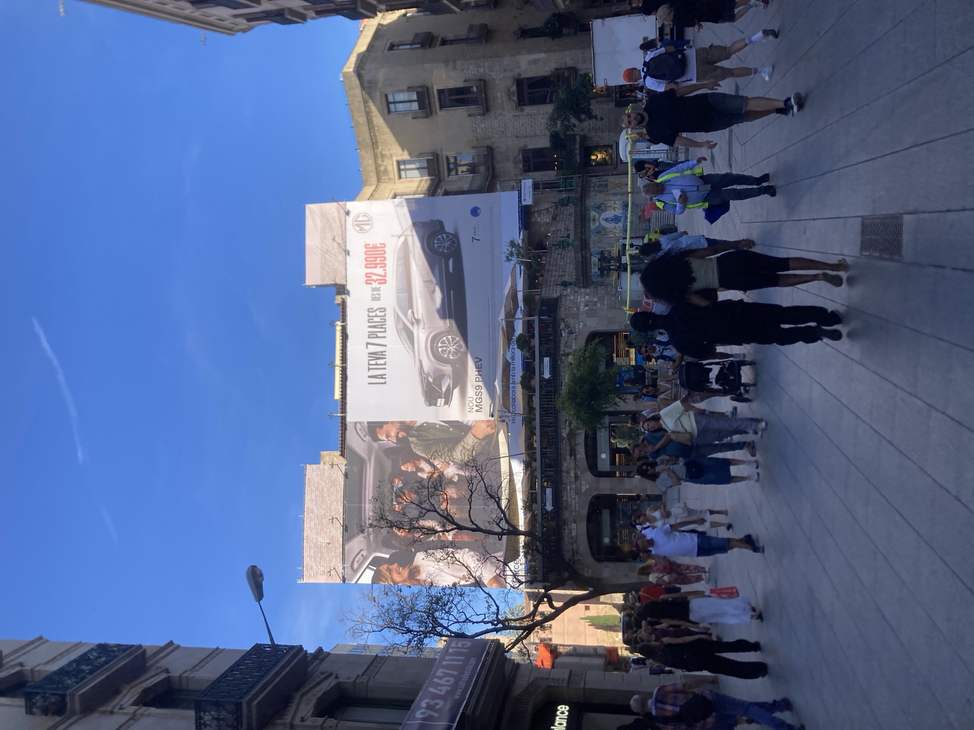 The image captures a street scene in Barcelona, Spain, likely in a commercial or pedestrian zone. Dominating the upper portion of the frame is a large billboard advertising the MG MGS 9 PHEV, highlighting "7 PLACES" and a price of "32.990€". The billboard features an image of the car and a family inside it. Below the billboard, the architecture of the buildings is visible, with balconies and arched entrances.
In the foreground, a crowd of people are walking along a wide, paved pedestrian walkway. They are dressed in casual summer clothing. Several individuals are walking towards the camera, while others are moving away. The atmosphere appears to be lively, with people engaged in everyday activities. A street lamp is visible on the left side of the image, and trees with bare branches are interspersed throughout the scene, suggesting a possible autumn or winter season, or perhaps they are deciduous trees that have been pruned. The sky is clear and blue, indicating good weather. The overall impression is that of a busy urban environment.