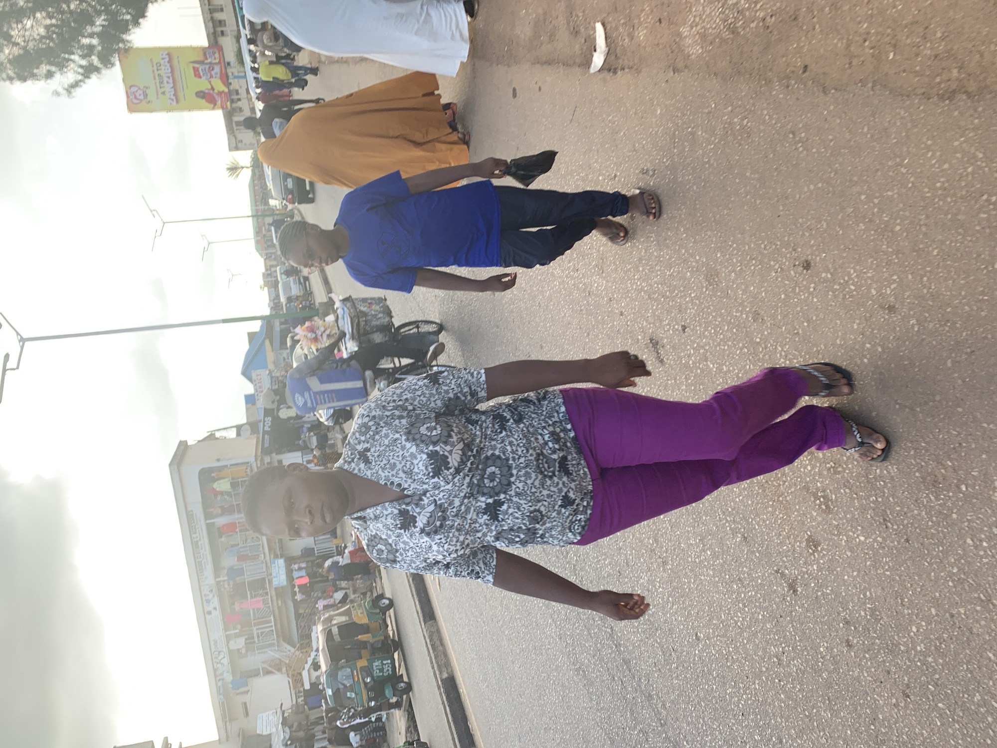 The image depicts a street scene in Jos, Nigeria, likely in a busy market area. In the foreground, a woman with short dark hair walks towards the viewer, wearing a black and white patterned blouse and vibrant purple trousers. Her right arm is bent at the elbow, and her left arm is extended slightly. Beside her, walking in the same direction, a young person wearing glasses, a blue t-shirt, and dark trousers, carries a black plastic bag in their right hand.
In the background, various elements of the urban environment are visible. A green and yellow auto-rickshaw is parked on the left side of the road. Several buildings line the street, some with awnings and displaying clothing on mannequins. Further back, people are walking and cars are present, indicating ongoing activity. A large yellow billboard advertising a trip to Zanzibar is visible on the right side of the frame. The sky is overcast with some sunlight filtering through, suggesting it might be late afternoon. The road surface is asphalt, with some debris visible. The overall impression is of a bustling commercial street with pedestrian and vehicular traffic.