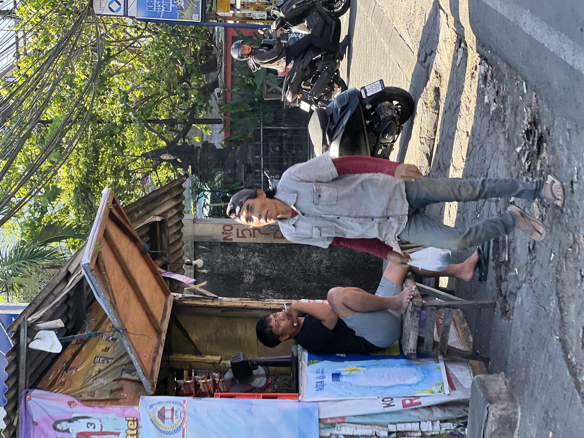 A street scene in Kuta, Indonesia, features two men and a motorcycle. A man in a black t-shirt and grey shorts sits on a wooden stool outside a small stall, smoking a cigarette. He is barefoot. Another man, wearing a grey button-up shirt with a red undershirt and jeans, walks past, carrying a white plastic bag. He wears a baseball cap and sandals. Behind them, a person in a black hoodie and helmet rides a black scooter. The stall has a corrugated metal roof and displays various posters and products, including a large blue banner advertising water. The ground is paved with rough asphalt and scattered debris. Lush green trees and overhead wires are visible in the background.