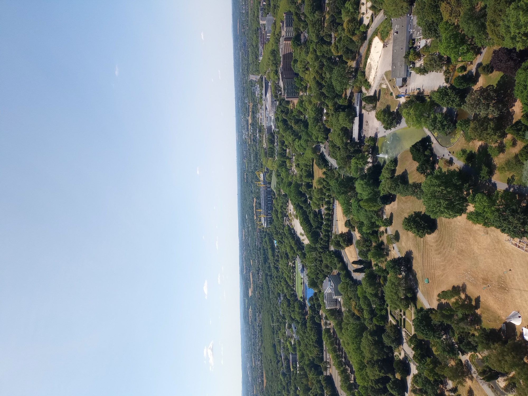 This is an aerial, wide-angle, high-definition photograph taken on a clear day overlooking a sprawling urban landscape interspersed with abundant greenery in Dortmund, Germany. The image captures a vast expanse of parks, trees, and modern infrastructure, including buildings and a large stadium, which is likely Signal Iduna Park, home of Borussia Dortmund.
In the foreground, a dry, grassy field is visible, with a few small tents and a wooden structure. Further back, a park area features a pond with a fountain and several pathways. A light-colored building with a flat roof, possibly a visitor center or restaurant, is situated next to the pond.
The mid-ground is dominated by dense deciduous trees, through which glimpses of roads and buildings can be seen. A large, bright blue swimming pool or pond is also visible among the trees. In the distance, a massive stadium with yellow and black accents stands prominently, its distinctive architecture identifiable. Several other large buildings and complexes are scattered throughout the cityscape, some appearing to be office buildings or educational institutions.
The background reveals a hazy horizon, with more urban development and rolling hills stretching into the distance. The sky is a clear, pale blue with a few small, wispy white clouds. The overall impression is of a well-integrated urban environment where nature and modern development coexist. No people are clearly discernible in the image.