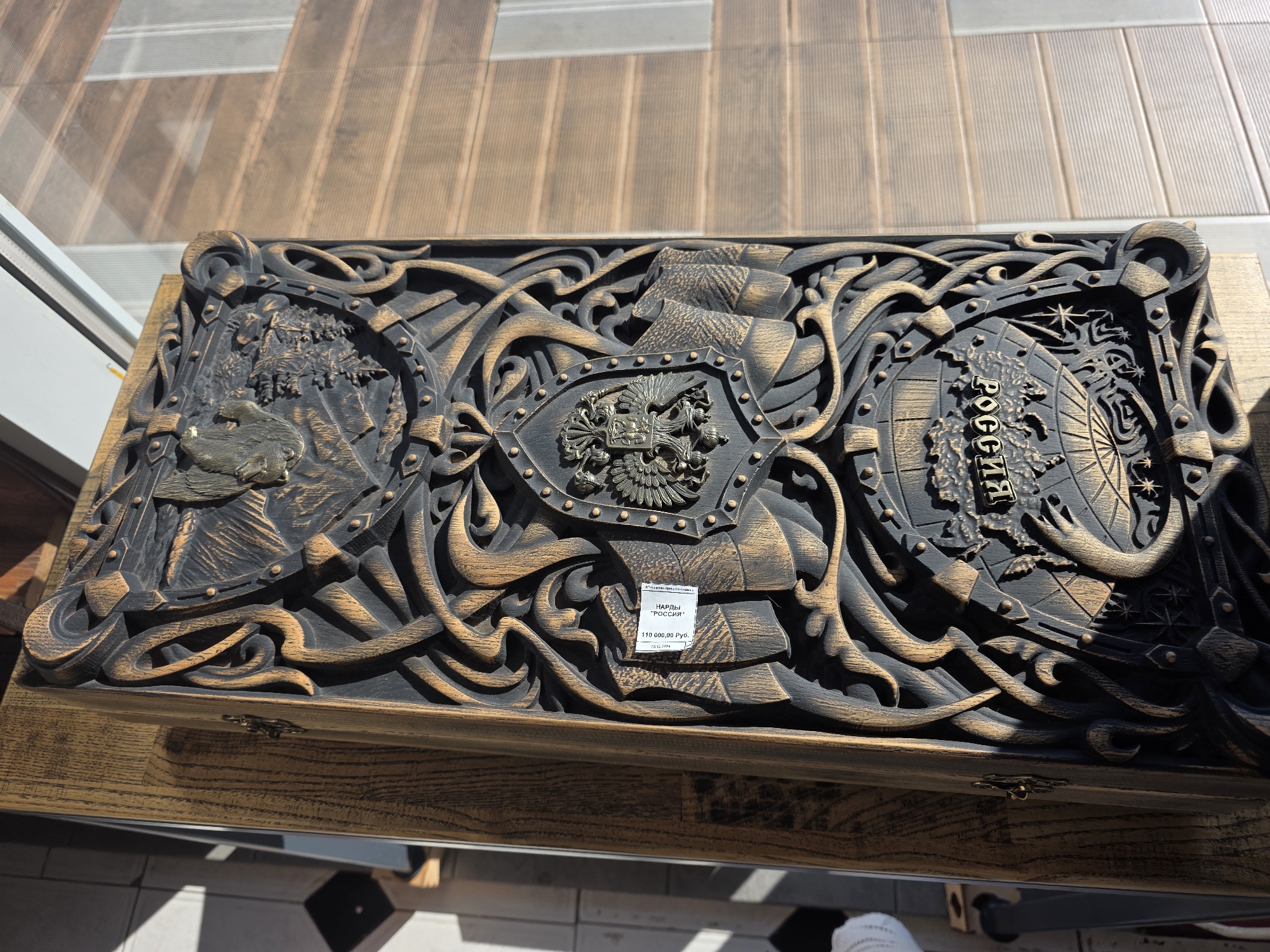 The image shows a detailed carved wooden object, possibly a box or a decorative panel, placed on a wooden surface. The carving features intricate designs, including what appear to be coats of arms, banners, and foliage. In the center of the object, there is a price tag attached that reads "110 000,00 RUB" and "РОССИЯ". The object is illuminated by sunlight, casting shadows and highlighting the texture of the carving. The background consists of a wooden wall or structure, and in the upper right corner, a window with horizontal blinds is visible. The overall impression is that of a display in a shop or a private collection.