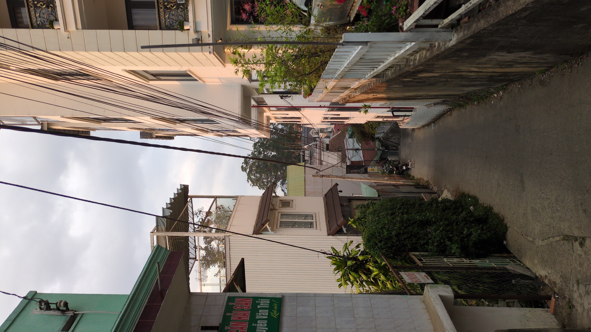 The image depicts a narrow street in Da Lat, Vietnam. The street is paved and runs between buildings of varying heights and architectural styles. Overhead, numerous electrical wires crisscross the sky. Buildings line both sides of the street, with some showing signs of aging. Vegetation is present, with trees visible in the distance and bushes nearer the camera. A few parked motorbikes are seen further down the street. The sky is overcast.