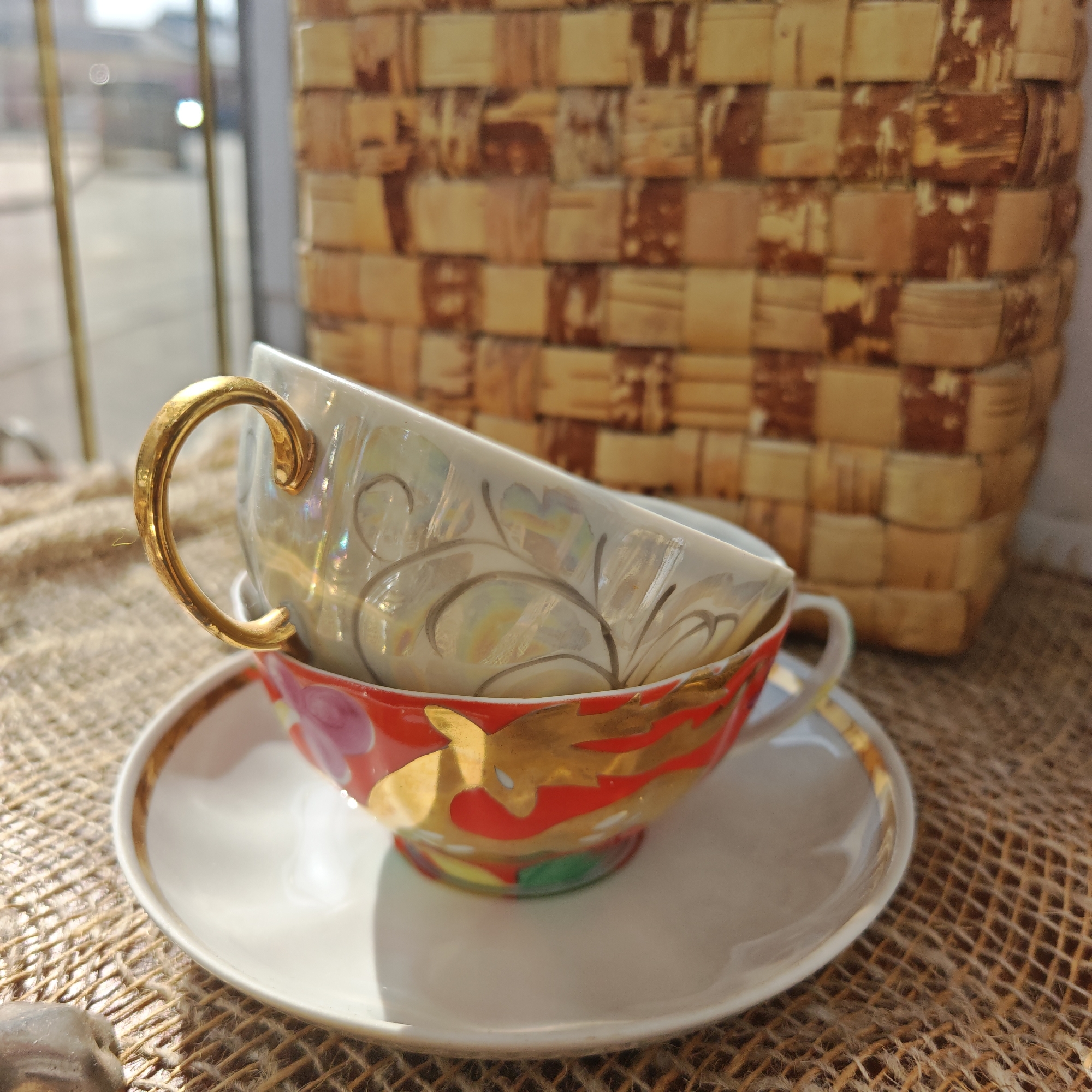 Two stacked tea cups and a saucer are positioned on a burlap-like surface, with a woven basket in the background. The lower cup is red with gold designs and rests on a white saucer with a gold rim. The upper cup is iridescent with a gold handle and grey filigree. Light from a window illuminates the scene, casting shadows. The window reveals an indistinct urban landscape outside. The location may be Rostov, Russia, as specified.