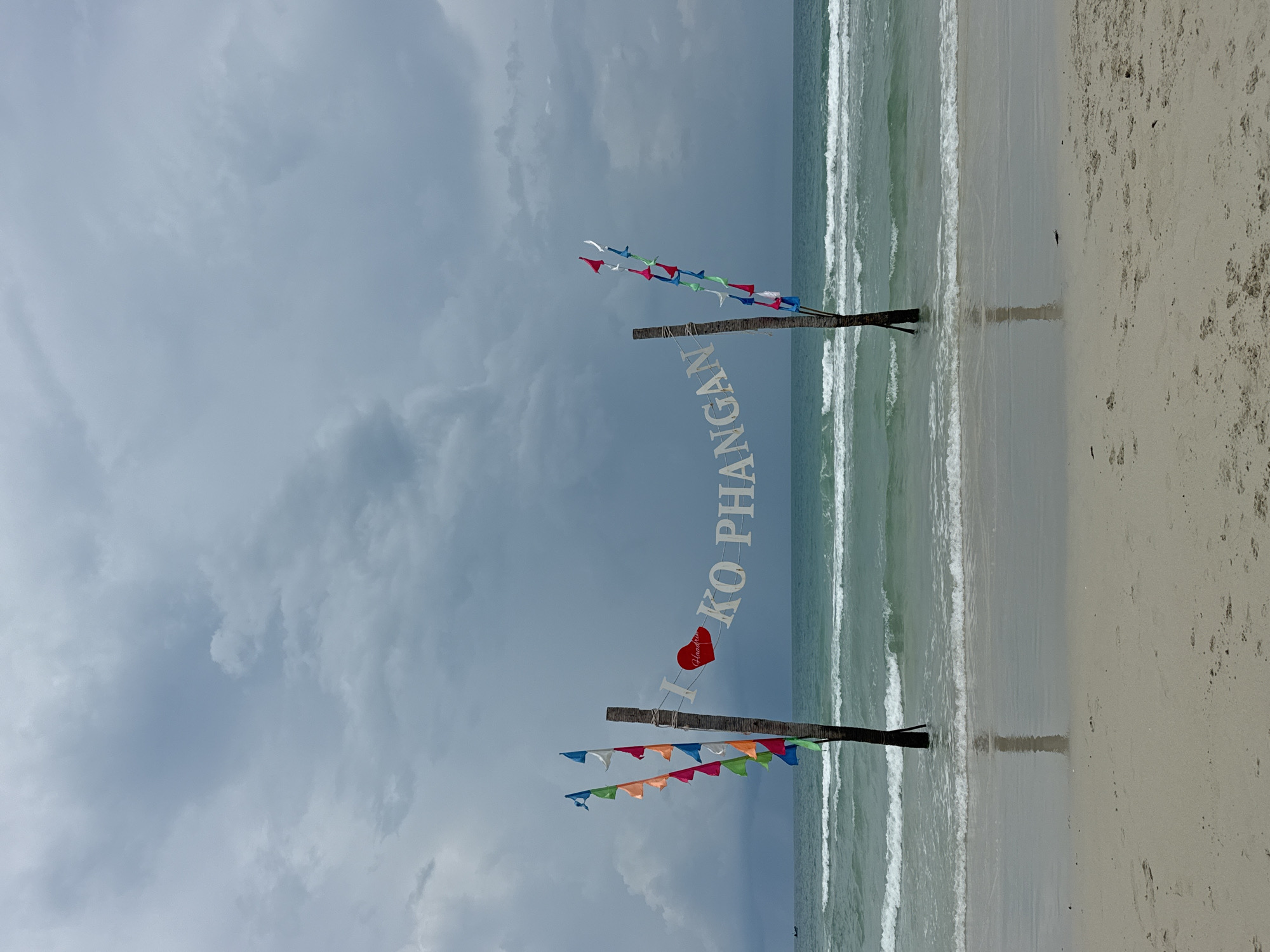 The photograph captures a scene at Ban Haad Rin, Thailand. A beach is present, with waves breaking on the sandy shore. Two wooden poles, each adorned with colorful triangular flags, stand on the beach. Between the poles, vertically oriented white letters spell out "I (heart symbol) KO PHANGAN." The heart symbol is rendered in red. The sky is overcast with cloudy conditions.