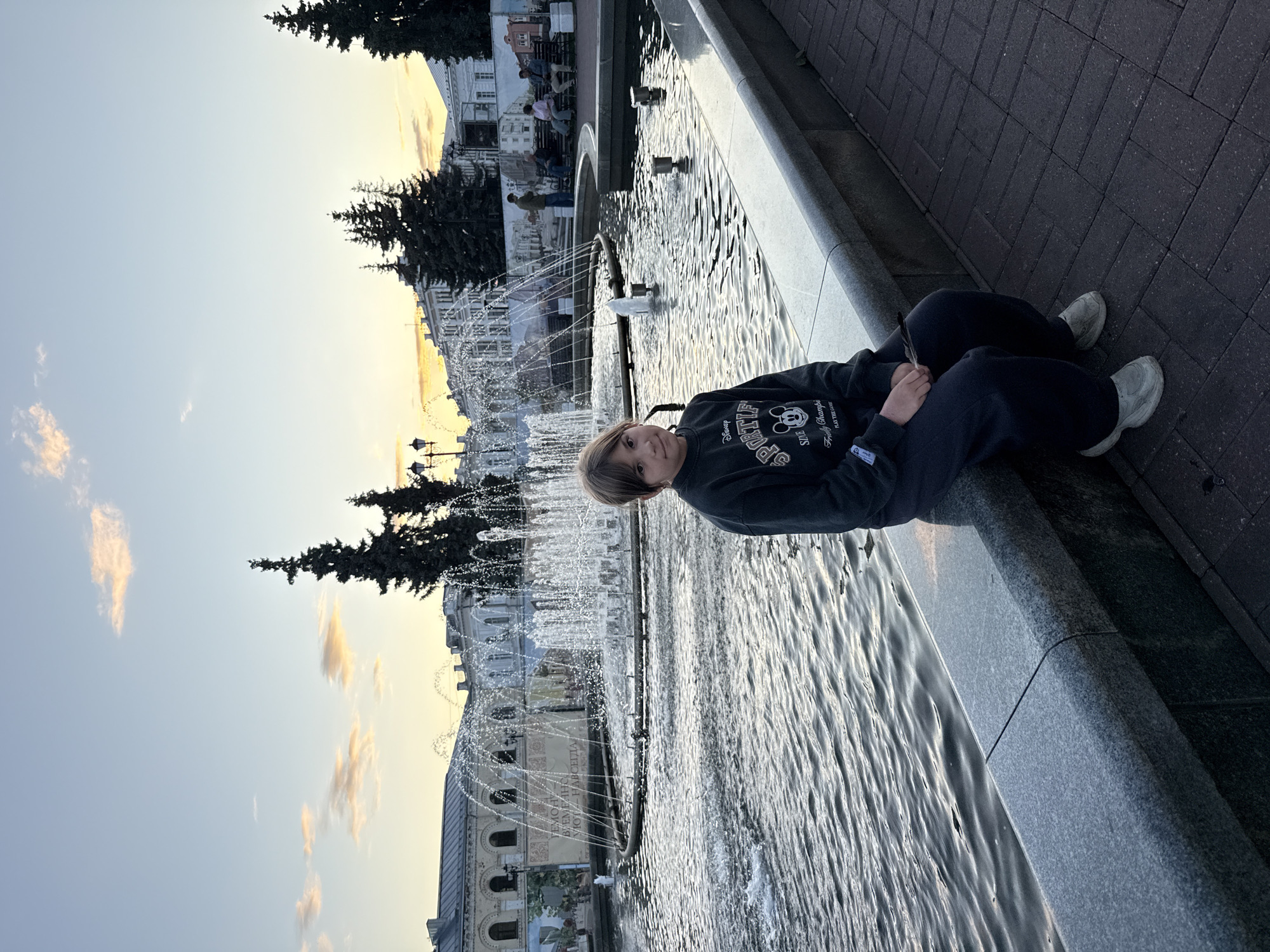 A young person sits on the edge of a stone-bordered fountain in a city square, likely in Rusanovo, Russia, given the context. The person is wearing a dark sweatshirt and pants, with light-colored shoes. Their hands are in their lap. The fountain has multiple jets of water spraying upwards, creating a shimmering effect on the water's surface. Behind the fountain, a row of buildings with classical architecture lines the square. Trees are visible interspersed between the buildings. The sky above is a soft gradient of blue and yellow, with a few wispy clouds, suggesting it is either sunrise or sunset. The ground in front of the fountain is paved with dark brick. The overall scene conveys a peaceful, urban atmosphere during twilight.