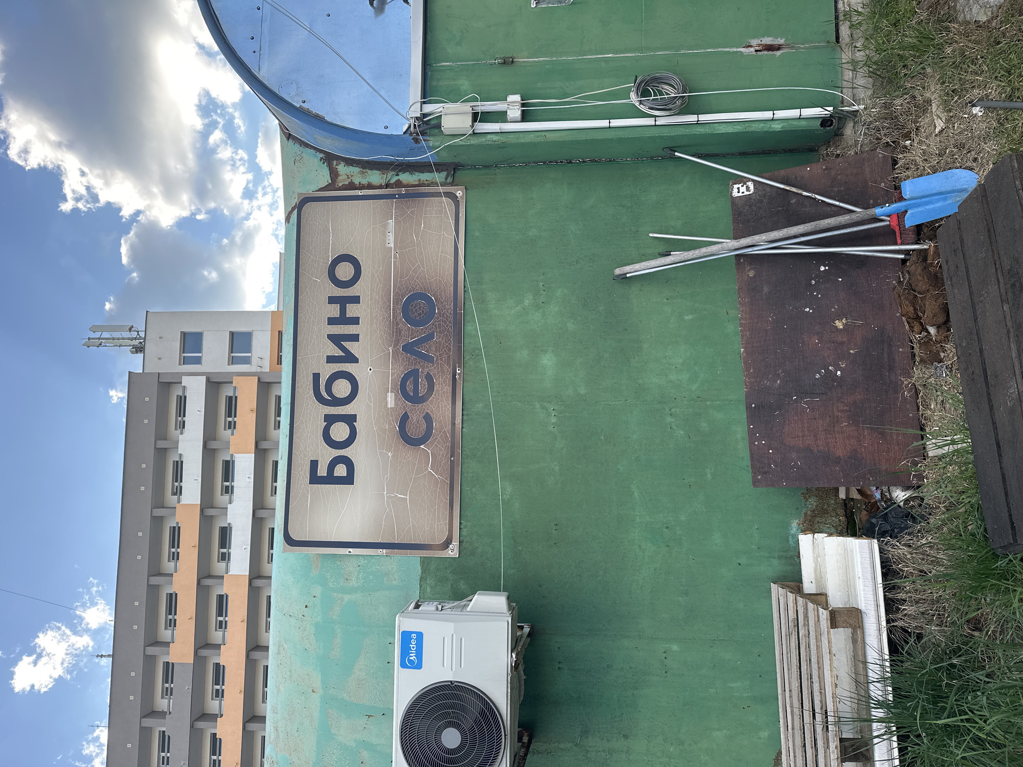The image shows an outdoor scene, likely a street view, in Sofia, Bulgaria. A weathered sign in Cyrillic script reads "Бабино Село" (Babino Selo) is mounted on a green exterior wall. Below the sign, an Midea air conditioning unit is attached to the wall. Adjacent to the wall, there's a collection of discarded items including a blue shovel, metal poles, wooden boards, and overgrown grass. In the background, a tall residential building with a mix of gray, white, and orange panels is visible under a blue sky with scattered clouds.