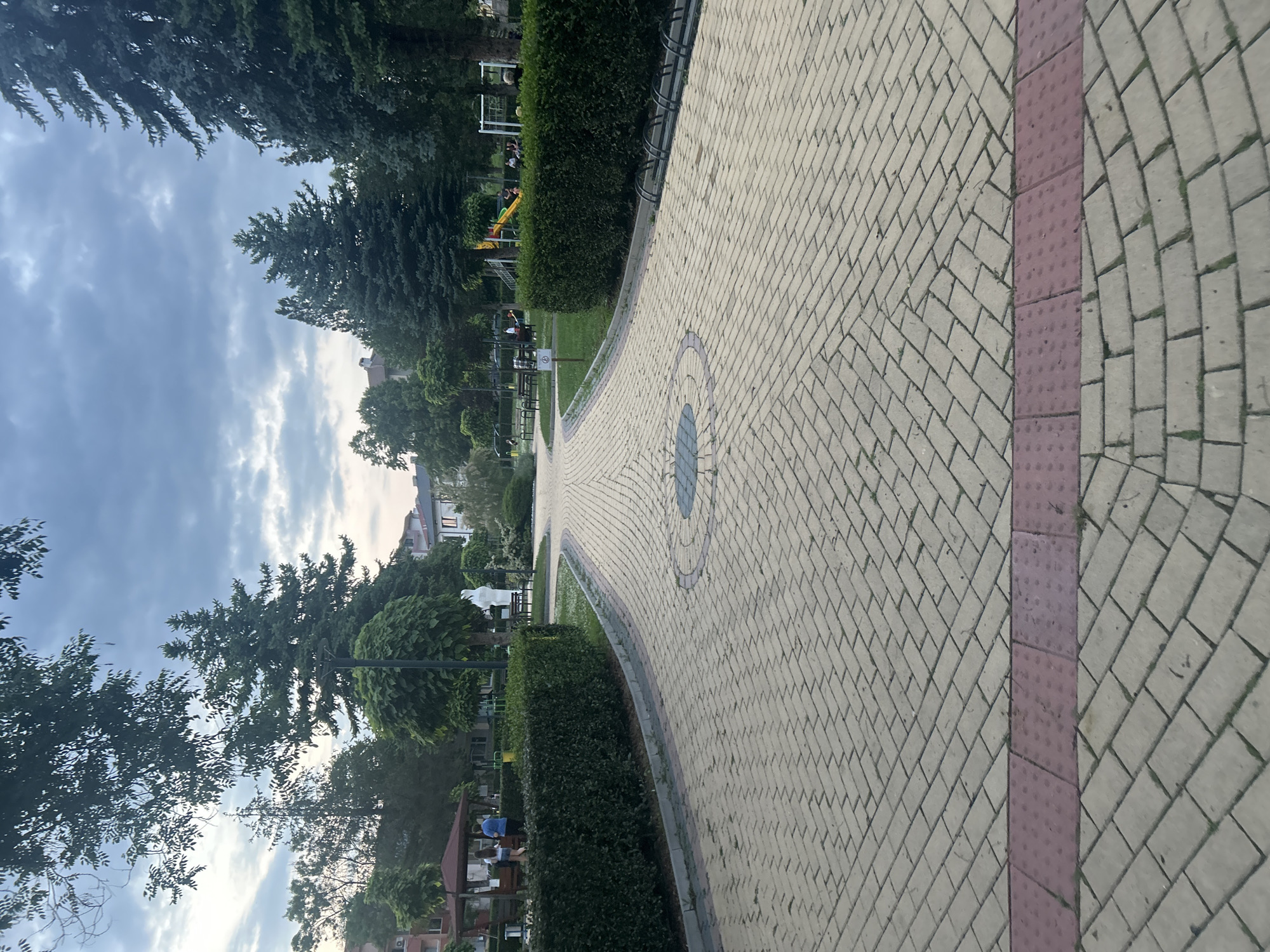 The image depicts a serene public park or garden in Dzhebel, Bulgaria, seen along a gently curving paved pathway. The path is constructed from light-colored rectangular pavers laid in a herringbone pattern, featuring a circular decorative inlay of darker blue and light pavers in the mid-ground. Along the right edge of the path, there is a continuous strip of reddish-purple textured tactile paving.
Lush green hedges line the pathway, separating it from grassy areas and numerous mature trees, including tall conifers and various broadleaf trees that frame the sky. In the distance, beyond the foliage, residential-style buildings are visible, suggesting an urban or semi-urban setting.
Several people are present, though mostly indistinct. On the left side, behind the hedge, a children's playground with yellow and red equipment is visible. Further along the path, individuals are seated on benches, one group under a wooden gazebo-like structure. Grey metal bicycle racks are installed along the path's edge near the benches. A white statue also stands amidst the greenery on the left.
The sky is partly cloudy with a soft, diffused light, indicating either late afternoon or early evening. The overall atmosphere appears peaceful and calm. A small white sign with an illegible symbol is faintly visible near a bench.