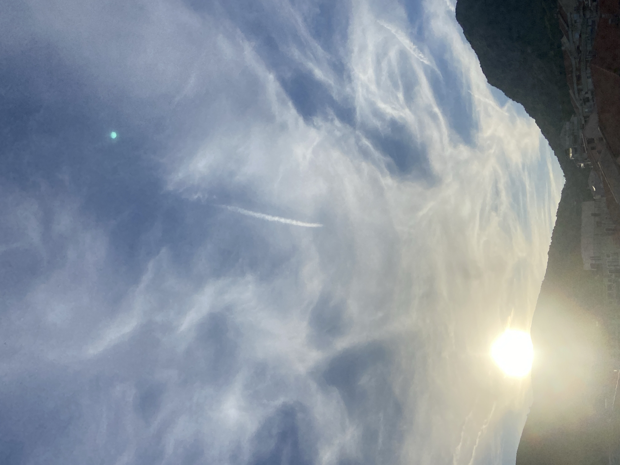 The image captures a sky over Budva, Montenegro, with wispy clouds and contrails from aircraft. The sun, a bright orb, is positioned on the left side of the frame, casting a warm glow over the hilly landscape and the buildings nestled within it. A dark, prominent mountain range dominates the right side of the image. The lower portion of the image reveals terracotta rooftops and some buildings, indicative of an urban setting. There are no people or active interactions visible in the scene. A small, bright cyan-colored circular anomaly is present in the upper right quadrant of the sky, possibly a lens flare or atmospheric phenomenon.