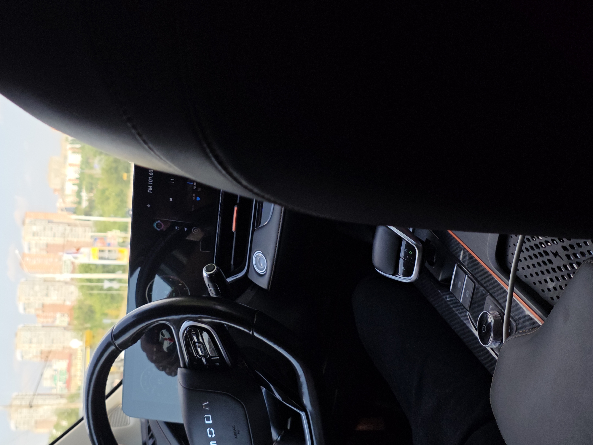 The image is an interior shot of a car's dashboard and front passenger seat, taken from the perspective of someone sitting in the back seat. The car appears to be in motion, as evidenced by the blurred buildings and trees visible through the windshield. The dashboard features a large, modern infotainment screen displaying "FM 101.60". To the left of the screen is a steering wheel with various controls. A gear shifter and a rotary dial with a power symbol are visible in the center console. The ambient lighting suggests it is daytime. The exterior view shows a cityscape with multi-story buildings under a bright sky, indicative of an urban environment. The specific location is identified as Rostov-on-Don, Russia.