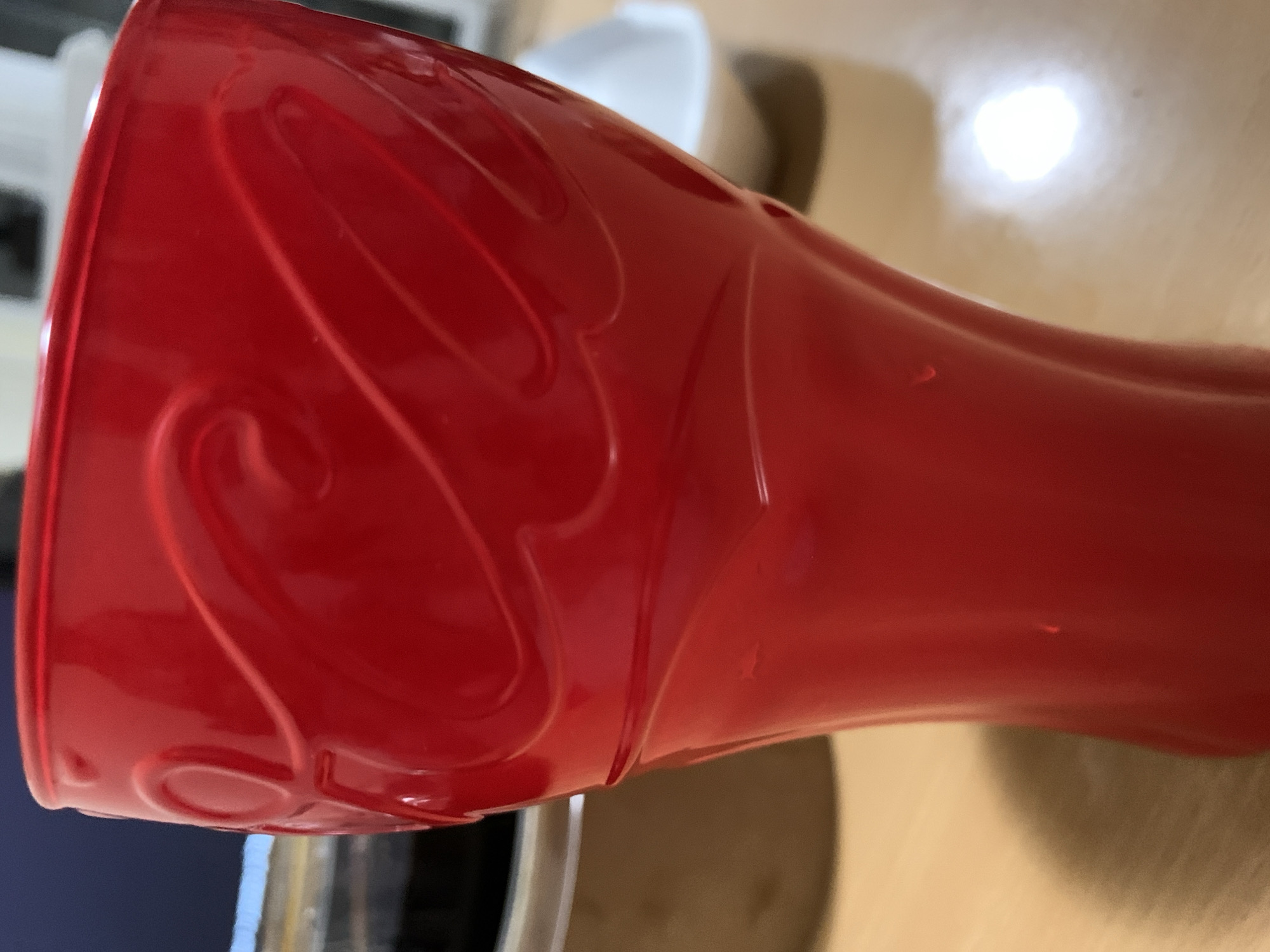 A close-up, slightly rotated, indoor shot focuses on a red, plastic, Coca-Cola cup. The cup has embossed detailing with curving lines and is positioned on a light-colored surface. Partially visible behind the cup are other objects, including a white bowl and what appears to be a kitchen sink area. There are no people visible. The scene lacks sufficient detail to determine a specific location in Sofia, Bulgaria.