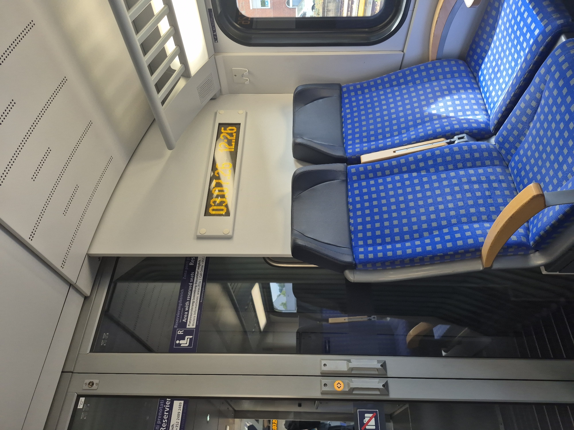 The image shows the interior of a train car with blue patterned seats. There are two seats visible in the foreground, with gray headrests and wooden armrests. A digital display above the seats shows the time "03:01:26 12:26". To the left of the seats is a glass door with a "Reserved Seats" sign. The environment appears to be clean and modern. The image is likely taken in Trier, Germany, as suggested by the information on the "Reserved Seats" sign, which mentions "Reservierung Luxemburg-Düsseldorf".
