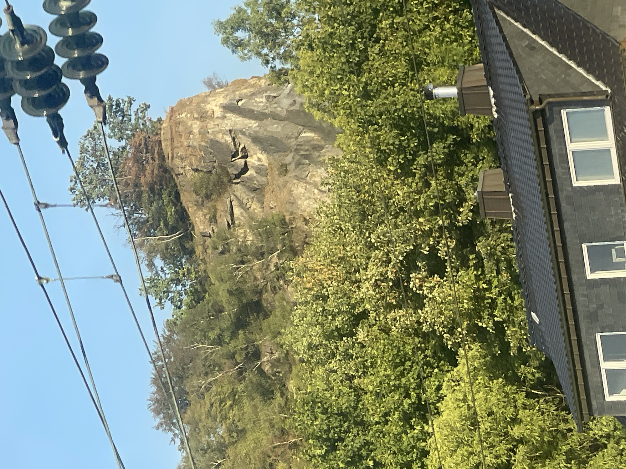 The image shows a rocky cliff face partially obscured by trees, with power lines strung across the foreground. In the lower portion of the image, the dark, tiled roof of a modern house is visible, featuring two chimneys, one of which has a silver chimney pipe. The scene is illuminated by natural daylight, suggesting it was taken outdoors. The location is identified as Iserlohn, Germany.