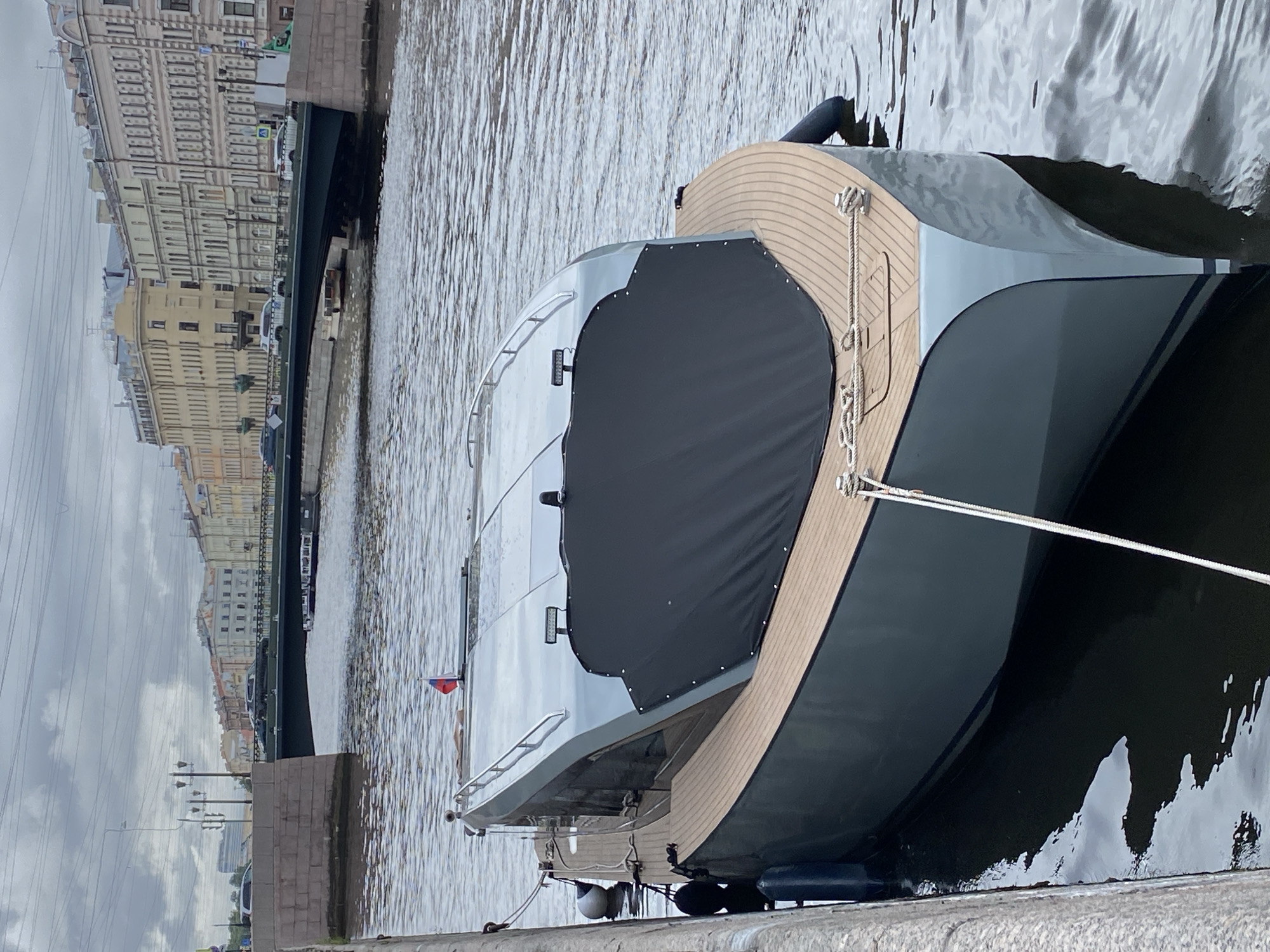 A modern, grey motor yacht is moored to a granite embankment on a cloudy day, likely in Saint Petersburg, Russia, given the architecture visible in the background. The yacht features a sleek design with a grey hull and a wooden deck at the bow. A black cover is draped over a portion of the deck. A small Russian flag flies from the stern. In the background, a bridge spans a waterway, with cars visible crossing it. The embankment is lined with old, multi-story buildings characteristic of St. Petersburg. The water is choppy, reflecting the overcast sky. There are no people visible in the image.