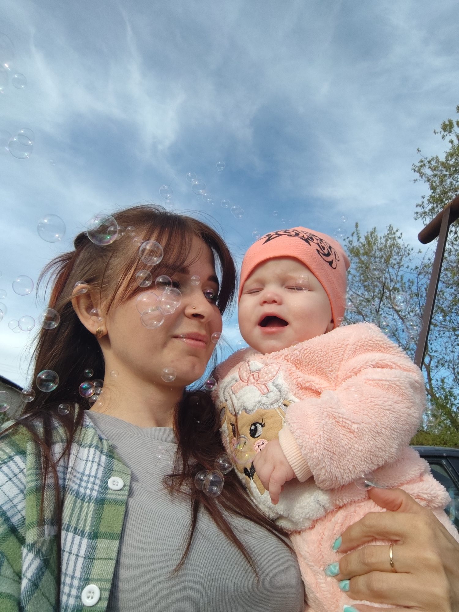 A woman holds a crying baby outdoors. The woman has brown hair and is wearing a green and white plaid shirt over a gray top. The baby is wearing a pink hat and a fuzzy pink outfit with a cartoon sheep design. The background is a blue sky with scattered clouds and glimpses of trees and a structure, possibly part of a swing set. Bubbles are visible in the air, creating a playful atmosphere. The woman's nails are painted in a light blue marble pattern. The image appears to be taken from a slightly low angle. Location: Фоки, Russia.