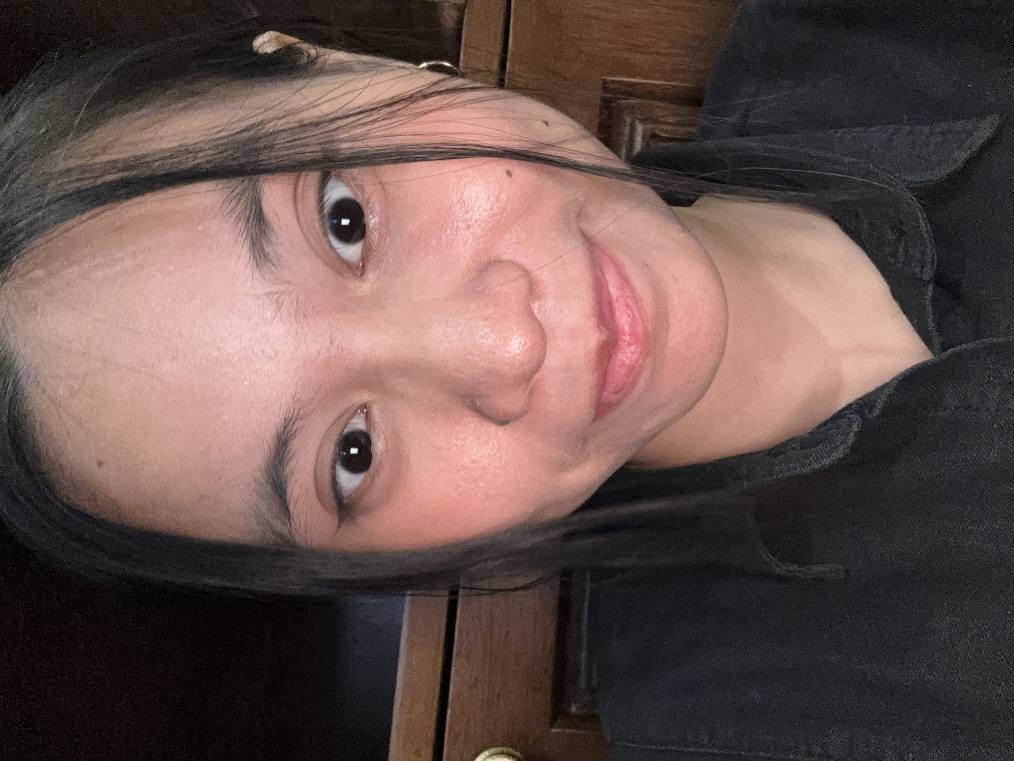 This is a close-up, indoor selfie of a young woman, likely taken in South Jakarta, Indonesia. She has long, dark hair, some of which falls across her face, framing her features. Her skin appears to be light-toned with visible pores and some redness on her cheeks, suggesting makeup application or a natural flush. She is wearing a dark-colored, collared shirt, possibly denim or a similar fabric, with a slight tear visible on the chest area. Her eyes are dark brown, and she is looking directly at the camera with a subtle smile, her lips slightly parted. A small hoop earring is visible in her right ear. The background is dark and somewhat out of focus, but appears to include wooden furniture, likely cabinetry. The lighting suggests an indoor environment, possibly a home or dimly lit room.