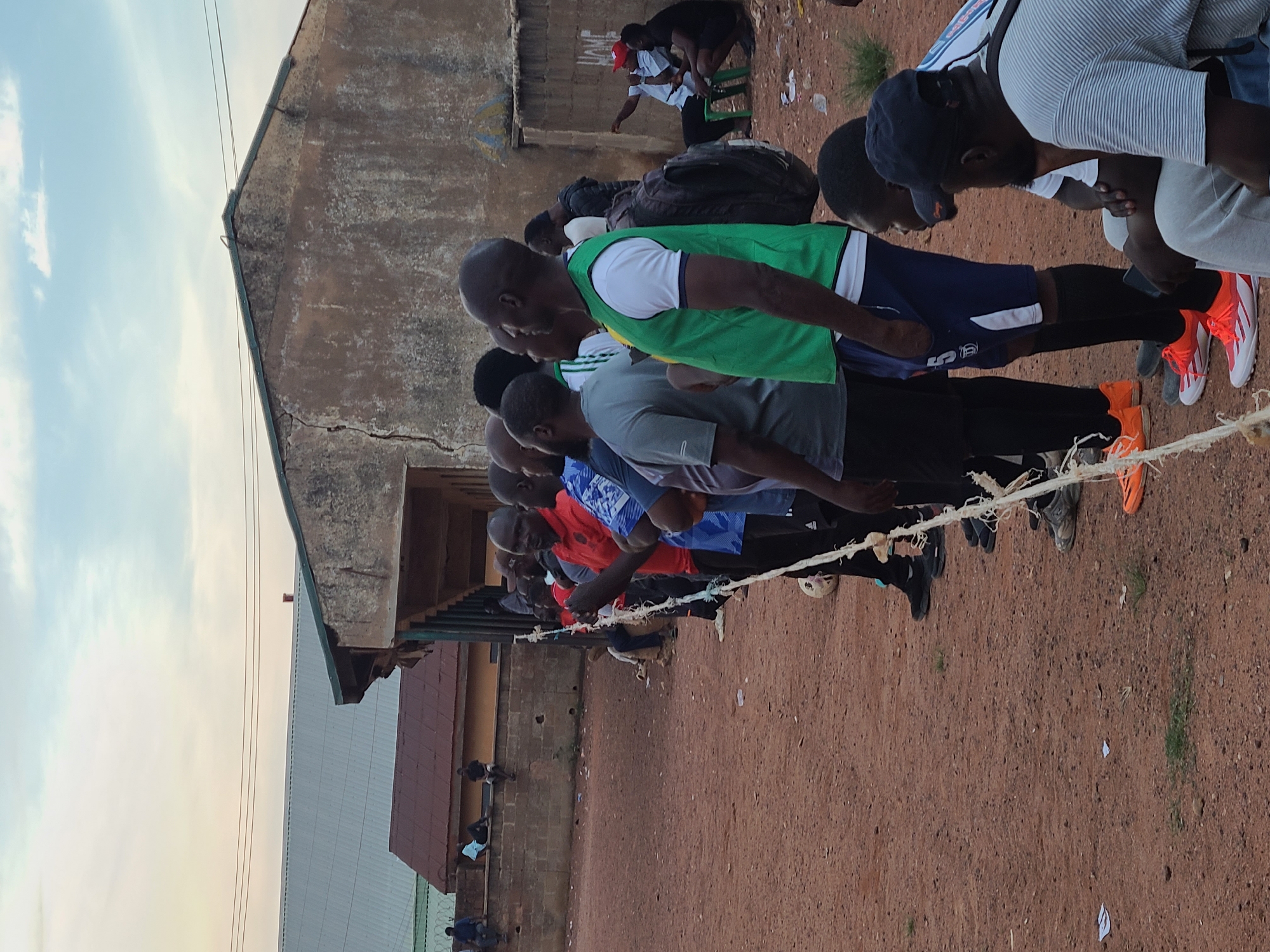 A group of men are gathered on a dirt field, possibly for a soccer game, in an urban setting in Jos, Nigeria. The sky is overcast with a light wash of pink and orange near the horizon. In the background, a dilapidated building with a cracked concrete wall and a corrugated metal roof stands prominently. A smaller building with a red metal roof is situated to the left, partially obscured by the main structure. Several men are standing in a line, facing away from the camera, with some wearing athletic attire. One man in the foreground, facing the camera, is wearing a red shirt and appears to be talking. Other individuals are seated on the ground or on low walls in the background, some watching the main group. The ground is a mix of dirt and scattered debris. A white rope, possibly marking a boundary, runs horizontally across the mid-ground of the image.