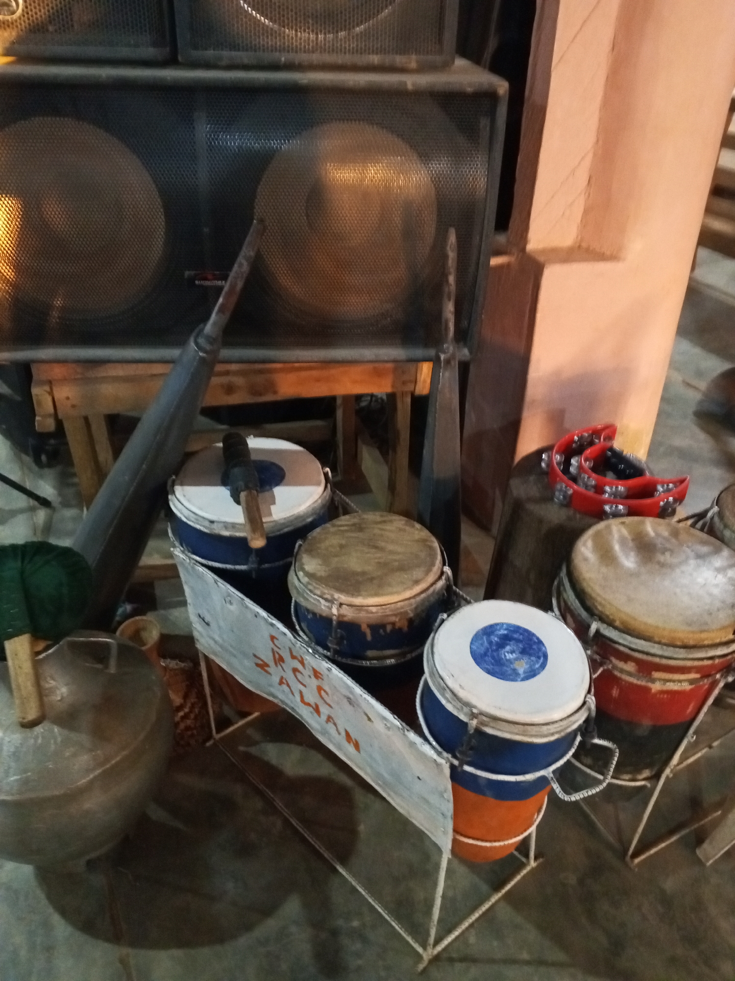 The image captures a collection of musical instruments, suggesting a practice or performance space in Unknown, Unknown. In the background, large speakers are stacked, and to the right, there's a peach-colored support structure. In the foreground, several drums of varying sizes and colors are grouped together, including a set with blue and orange accents. A tambourine rests on a small wooden stool. A metal pot with a green mallet sits to the left, along with a long metal pole. A sign with the text "CR C.F. ZAHAN AWAN" is visible near the drums. The scene is dimly lit.