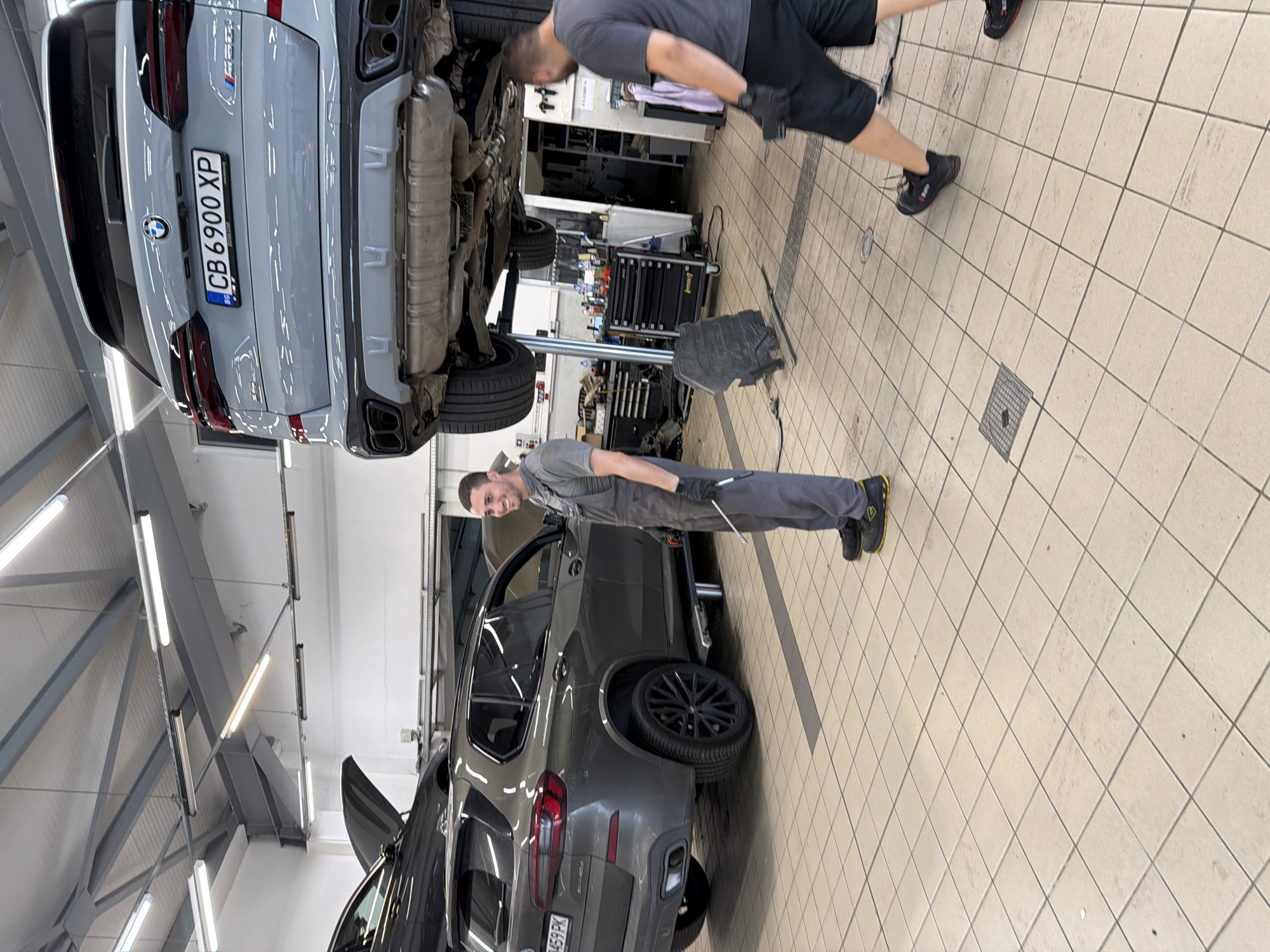 This is a photograph taken inside an automotive repair shop located in Burgas, Bulgaria. The scene features two men working on vehicles. In the foreground, a man wearing grey overalls and black gloves stands near a dark grey BMW SUV, holding a tool. This vehicle is on a hydraulic lift. In the background, another man wearing a dark t-shirt and black shorts is walking away from the camera. Partially visible in the background is the rear of a light grey BMW SUV, also on a lift, displaying the license plate "CB 6900 XP". The environment consists of a tiled floor, overhead fluorescent lighting, and various pieces of automotive equipment including tool cabinets and shelving.