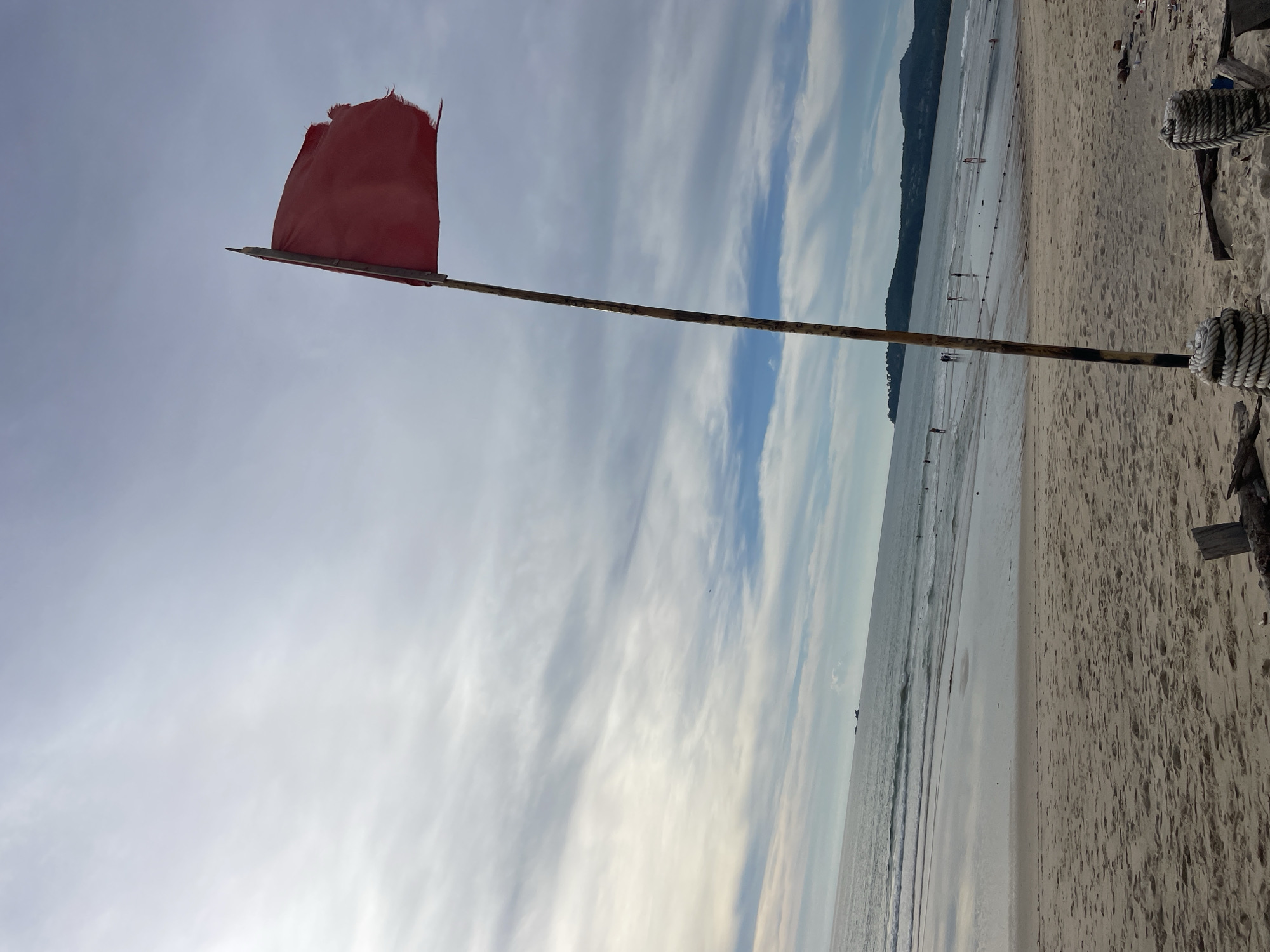 A red flag waves on a wooden pole on a sandy beach. The flag appears weathered and torn. The ocean is visible in the background with gentle waves and a few people scattered along the shore. A distant treed landmass forms the horizon. The sky is cloudy with streaks of light. The foreground shows sand, driftwood, and rope. The scene suggests a warning or a caution, possibly related to swimming conditions.