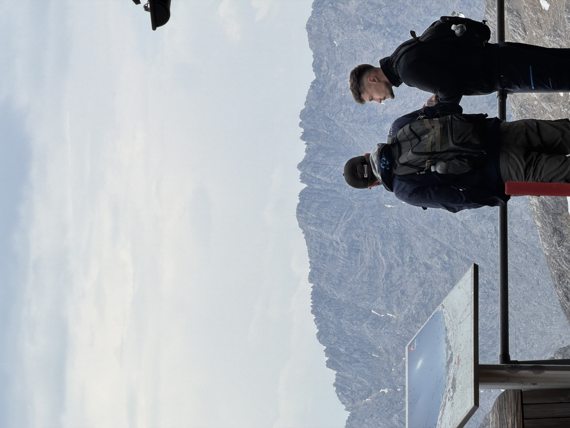 Two men are standing on a viewing platform, looking out at a mountain range under a cloudy sky. The man on the left has his back to the camera and is wearing a dark blue jacket, grey pants, and a dark baseball cap. He has a green backpack with a water bottle attached. The man on the right is facing left, looking towards the other man, and is wearing a black jacket and black pants. He has a black backpack with a water bottle attached. In the foreground, to the left, there is a signpost with a map displayed on it. The platform has a railing. The background consists of rugged, grey mountains with patches of snow and a pale, overcast sky. This scene is likely on the Zugspitze, the highest peak in Germany, located near Garmisch-Partenkirchen.