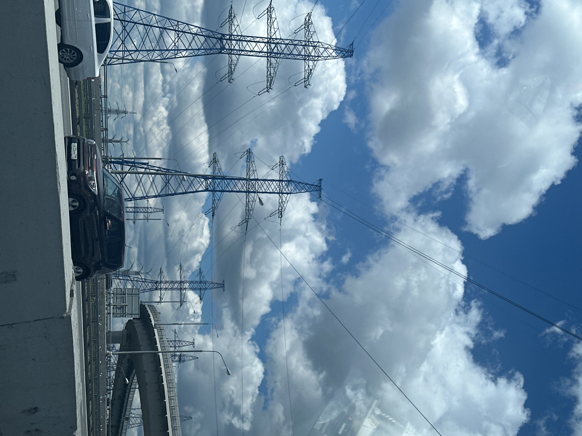 The image shows a view from a vehicle on a highway, with multiple high-voltage power line towers dominating the scene against a backdrop of a blue sky with scattered white clouds. In the foreground, a concrete barrier separates the viewer's lane from other lanes of traffic. A white sedan is visible in the left lane, and a dark-colored compact SUV with a license plate reading "c633km 777" is in the right lane. A highway overpass with a curved ramp is visible in the mid-ground. The overall scene suggests an industrial or semi-urban environment with extensive electrical infrastructure.
