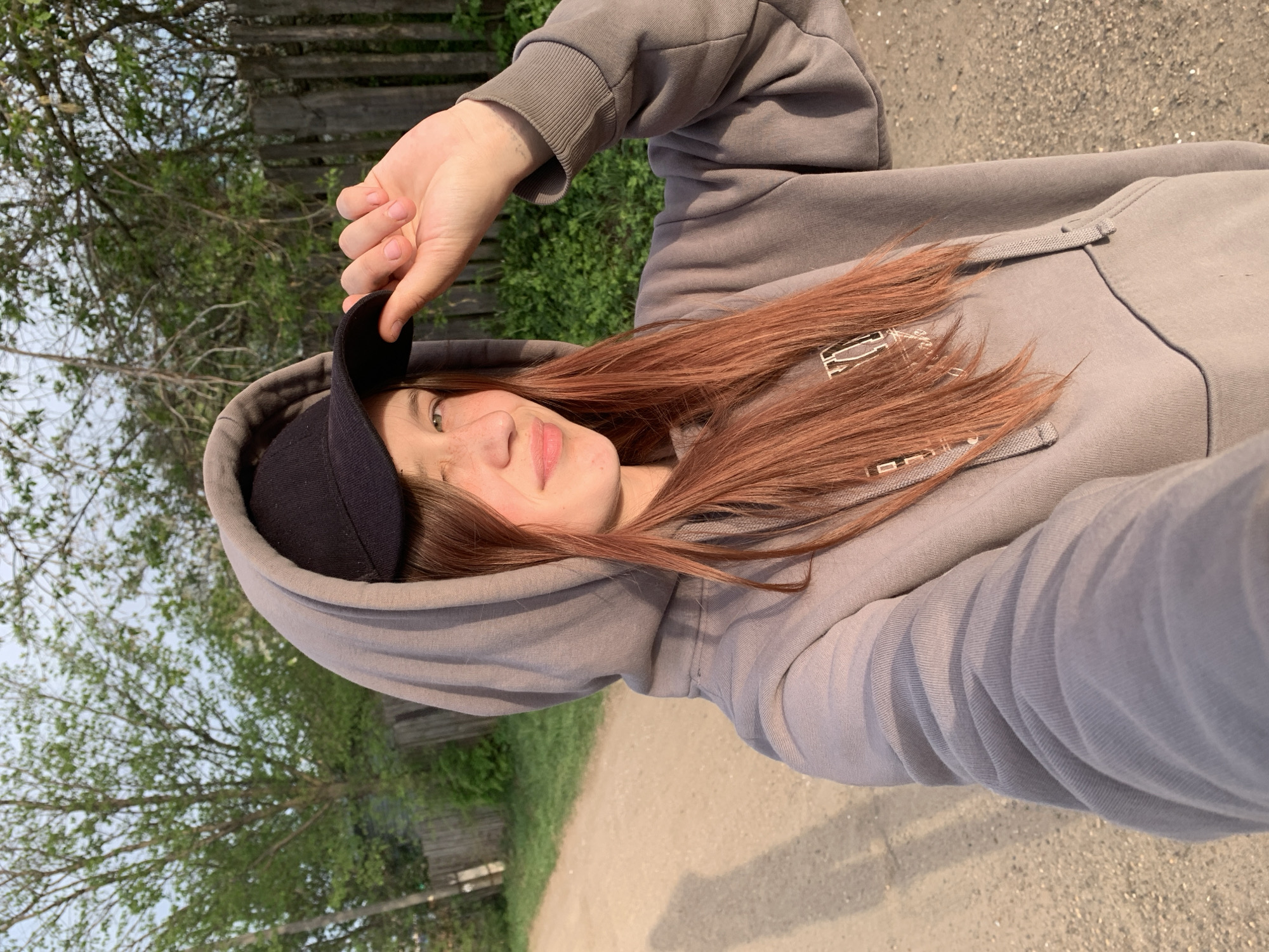 A young woman is seen in a selfie taken at ground level, possibly along a dirt path. She is wearing a gray hooded sweatshirt and holding the brim of a black baseball cap slightly elevated above her head. Her long auburn hair falls loosely around her shoulders. The background shows trees and greenery, with a wooden fence visible to the left. The dirt path is illuminated by sunlight. It is not possible to determine the exact location within the city of Фоки, Russia from the given information.