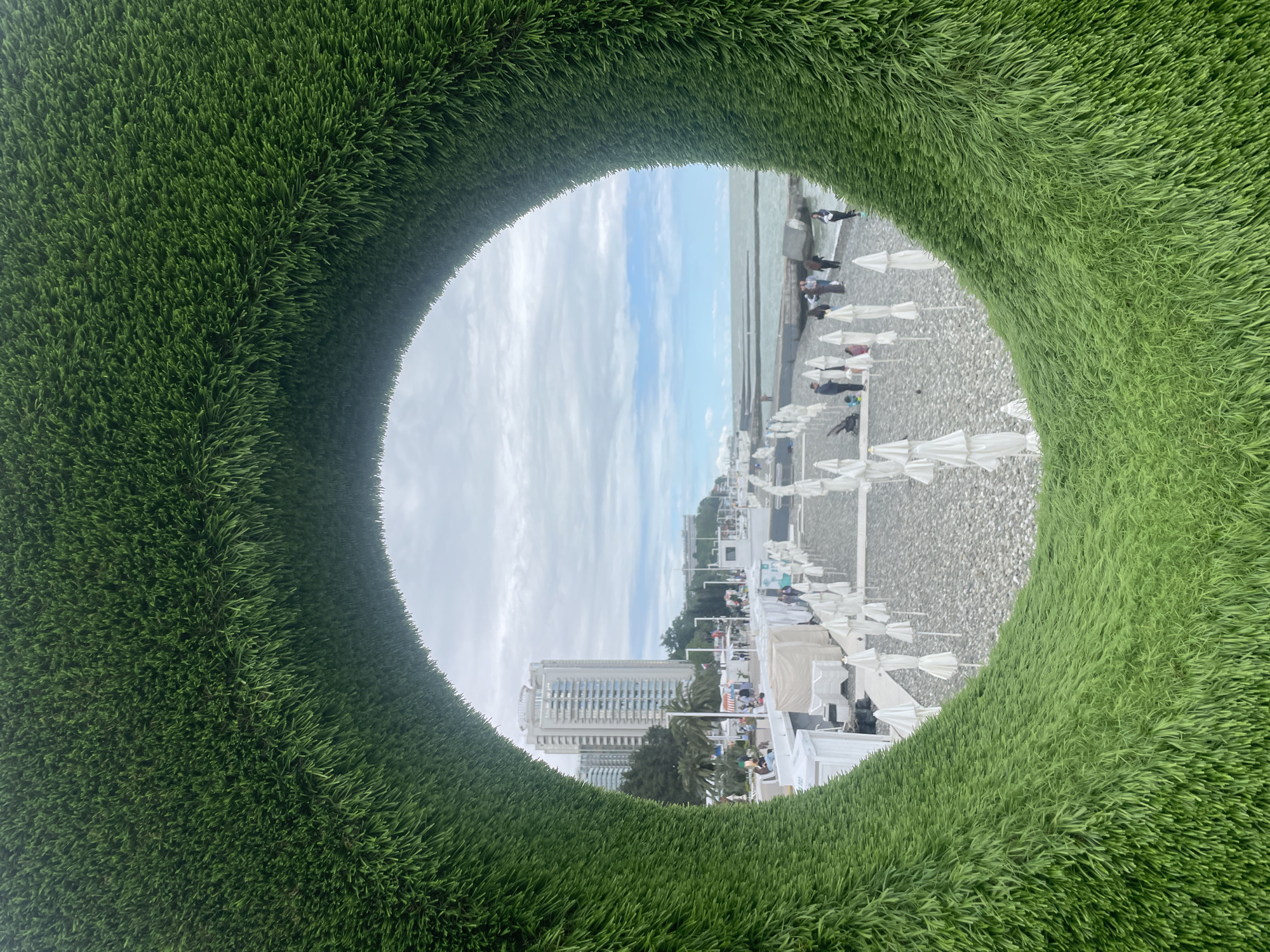 The image shows a view of a beach scene in Sochi, Russia, framed by a circular opening covered in artificial grass. The opening provides a unique perspective of the beach, where rows of white umbrellas and lounge chairs are set up on the pebble-covered ground. People are visible on the beach, some walking and others relaxing under the umbrellas. In the background, a tall building is visible, along with trees and a cloudy sky. The artificial grass surrounding the circular opening appears well-maintained and adds a contrasting element to the scene.