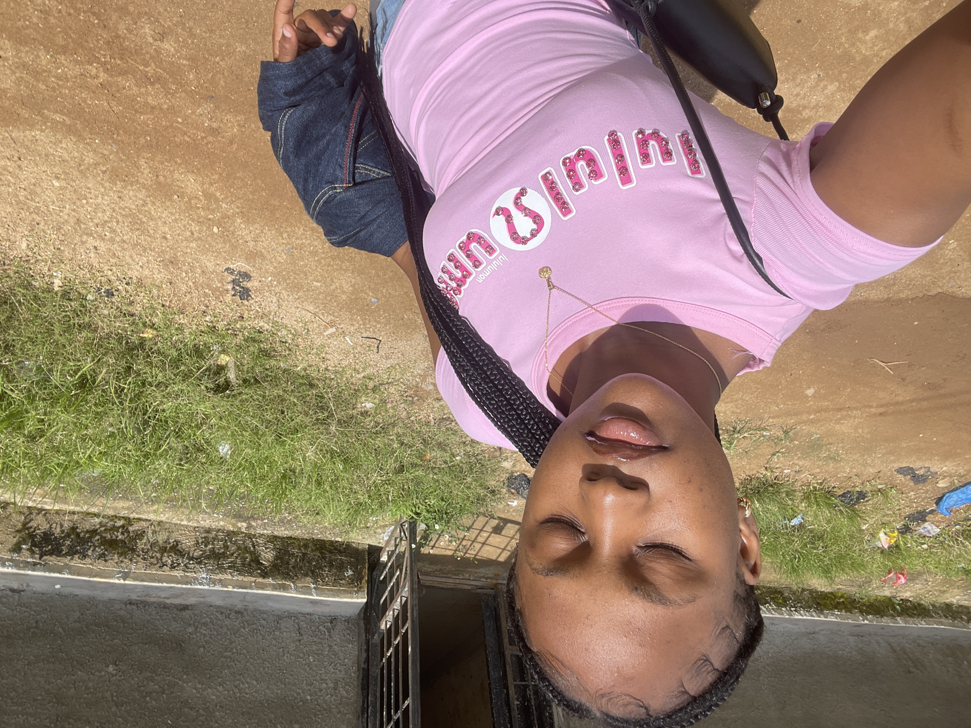 A young Black female, with braided hair and closed eyes, stands facing the camera outdoors in Rafin Par, Nigeria. She wears a pink t-shirt with a white and pink logo and a gold necklace. She holds a dark jacket over her right arm and a black purse is slung across her body. The ground is a mixture of dirt and gravel, with patches of green grass and some scattered debris. In the background, a building with a concrete wall and a metal gate is visible, with more grass growing around its base. The lighting suggests it is daytime.