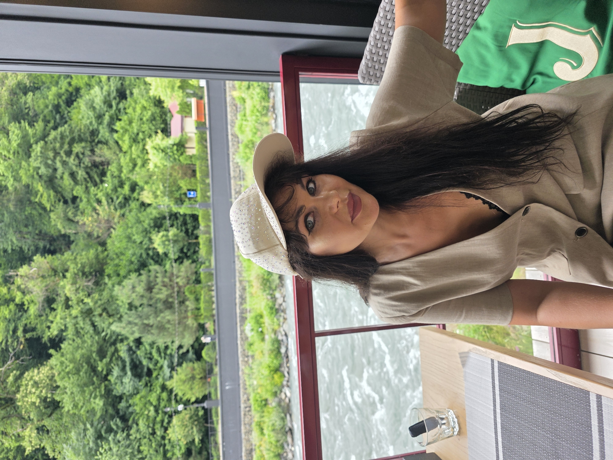A medium shot captures a woman sitting at a table on a covered patio, with a river and lush green mountains in the background. The woman, facing the camera, wears a beige cap adorned with rhinestones and a loose-fitting beige button-up shirt, revealing a black lace bra. Her long, dark hair is worn down, and she has dark eye makeup and burgundy lipstick. She is seated in a wicker chair next to a green cushion with a stylized "J" logo. On the wooden table in front of her, a glass with a dark object inside and a placemat are visible. The patio features a red railing. Beyond the railing, a river flows rapidly, and across the river, a tree-lined embankment with buildings and a road is situated at the base of densely forested mountains. The sky is overcast, suggesting a warm, possibly humid day. The location is identified as Estosadok, Russia.