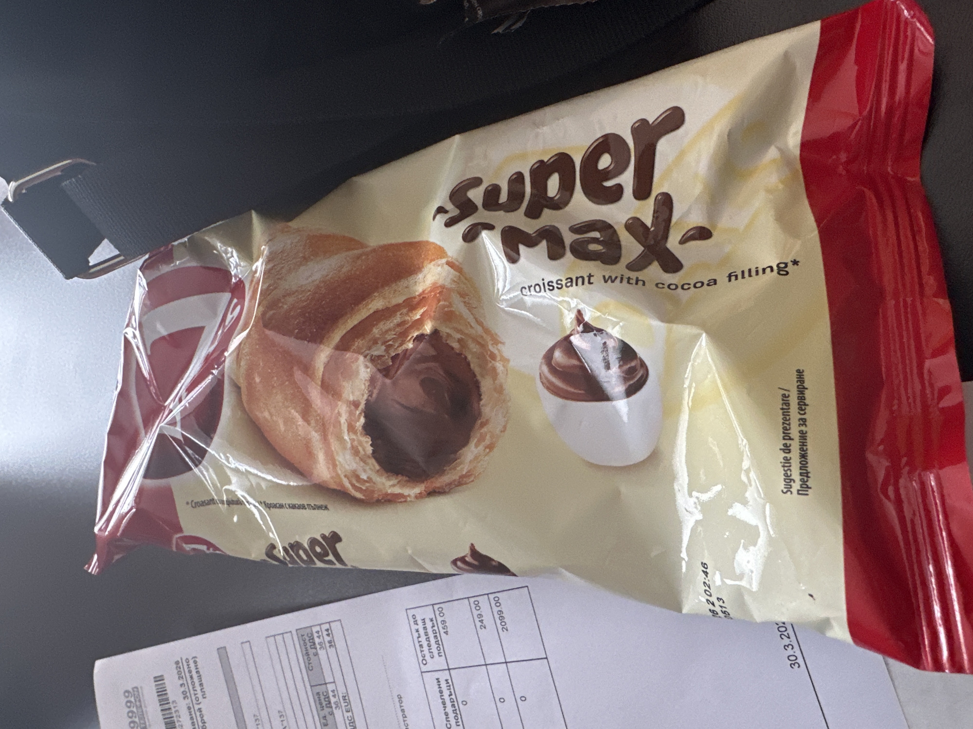 The image shows a package of "Super Max" croissant with cocoa filling. The package is predominantly white with brown lettering and a red strip on the right side. A croissant with visible chocolate filling is displayed on the package, along with an illustration of a scoop of chocolate. A sales receipt with Bulgarian text is beneath the package. The date on the receipt is 30.3.2026. While I cannot determine the location in Sofia, Bulgaria from the image, the Bulgarian text on the receipt suggests its location.