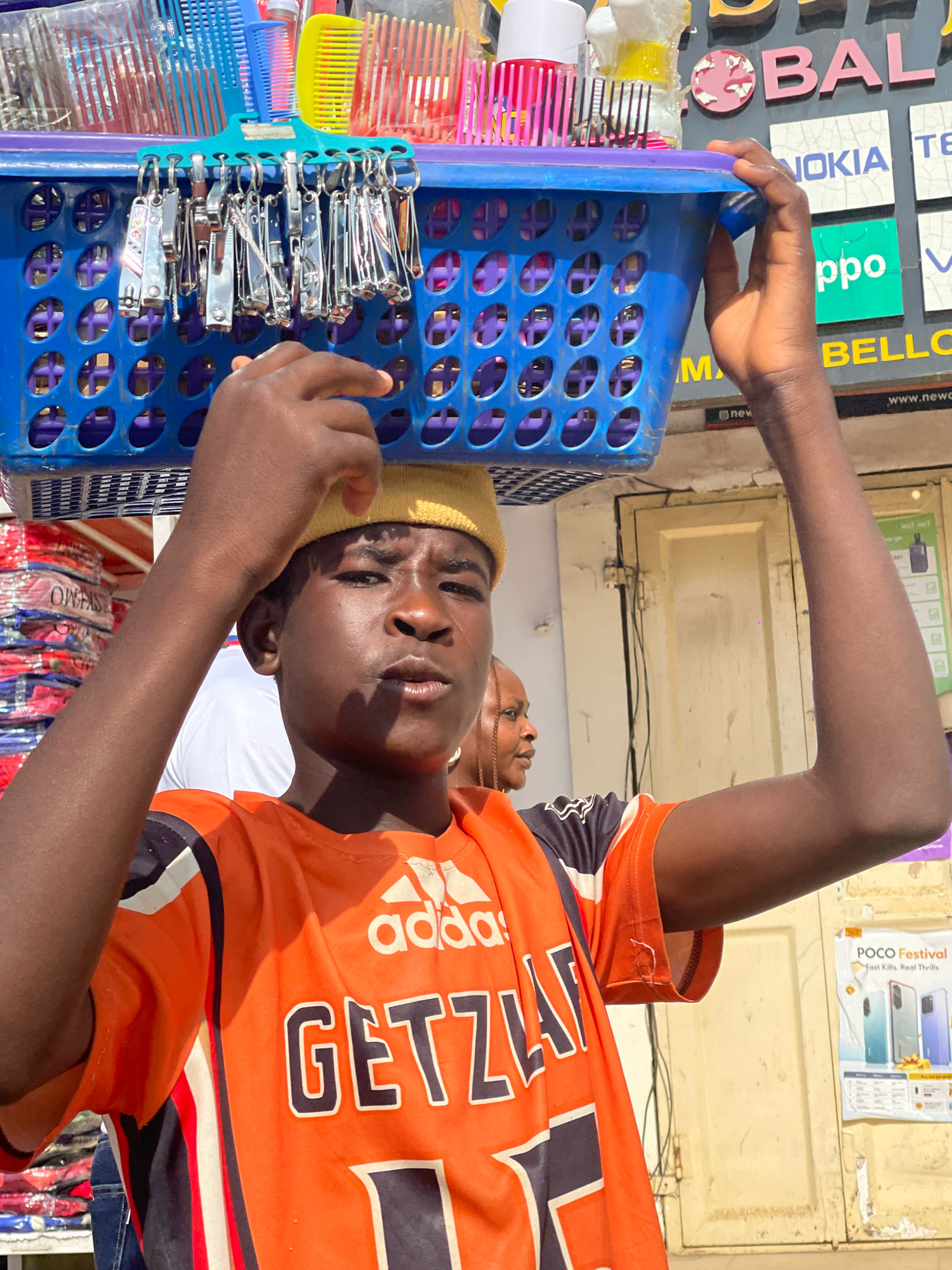 A young Black male wearing an orange Adidas t-shirt and a yellow beanie balances a blue plastic basket filled with various grooming tools on his head. The basket is adorned with numerous nail clippers, combs, and other small accessories hanging from its sides. In the background, a store facade is visible with signs for "GLOBAL", "NOKIA", "OPPO", and "BELLO". The scene appears to be in a bustling market or street setting in Jos, Nigeria. The young man is looking directly at the camera with a neutral expression. Another person is partially visible behind him to the right. The lighting suggests it is daytime.
