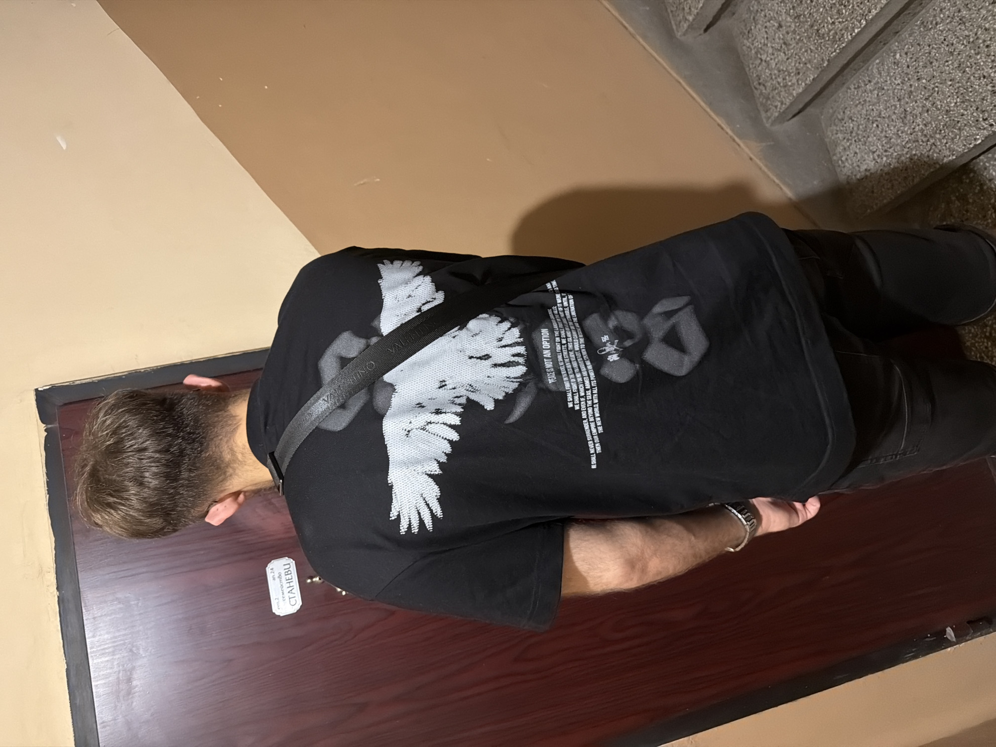 The image shows the back of a person standing in a narrow hallway or stairwell. The person is wearing a black t-shirt with a graphic print of a white bird with outstretched wings and text below it, and dark pants. A black strap, possibly from a bag, crosses the person's chest. The walls are painted a light, sandy color, and a dark wooden door is visible to the left. To the right, a concrete staircase with speckled treads is partially visible. A small white sticker with Cyrillic text, "СТАНЕВУ", is affixed to the door. The lighting appears to be artificial and casts shadows. The overall impression is an indoor residential or communal space. The location is not identifiable as Plovdiv, Bulgaria.