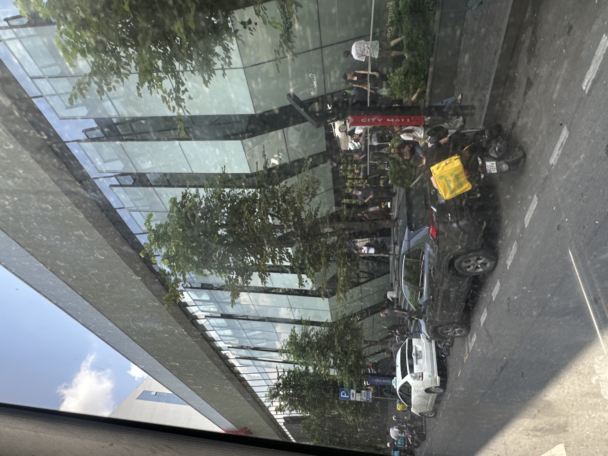 In Tbilisi, Georgia, a street scene unfolds in front of a modern building with a glass facade. The building's exterior reflects the blue sky and scattered clouds, with trees and architectural elements visible in the reflections. A grey overhang runs across the top of the building.
On the street, a white Toyota Prius, possibly a taxi, is parked. To its right, a dark-colored SUV is also parked, with its doors open and a person leaning against it. A motorcycle with a large yellow delivery bag is parked behind the SUV. The delivery bag has the "Glovo" logo visible.
A group of people are gathered near the entrance of the building, some standing and others sitting. Traffic signs, including a blue parking sign, are visible on the sidewalk. The street is paved with asphalt and marked with white lines. The overall impression is a busy urban environment on a sunny day.