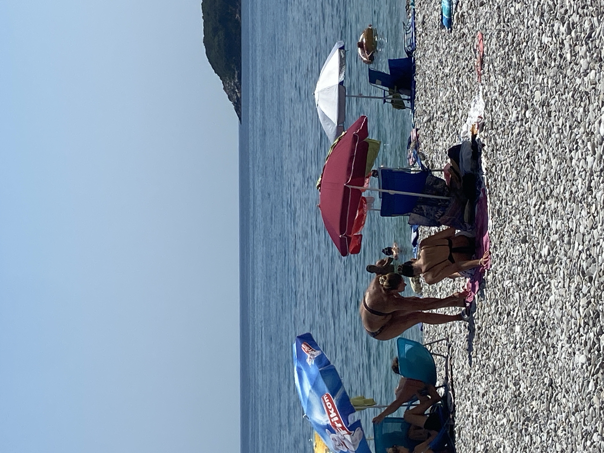 The image depicts a sunny day at a pebble beach in Prijevor, Montenegro. Several people are enjoying the water and relaxing on the shore. In the foreground, two women are on the beach, one bending down and the other seated. Various beach umbrellas and chairs are set up, providing shade. The sea is calm and blue, with a distant green, tree-covered island on the horizon. A person is visible floating on an inflatable ring in the water. The overall scene is one of leisure and a typical summer day at the beach.