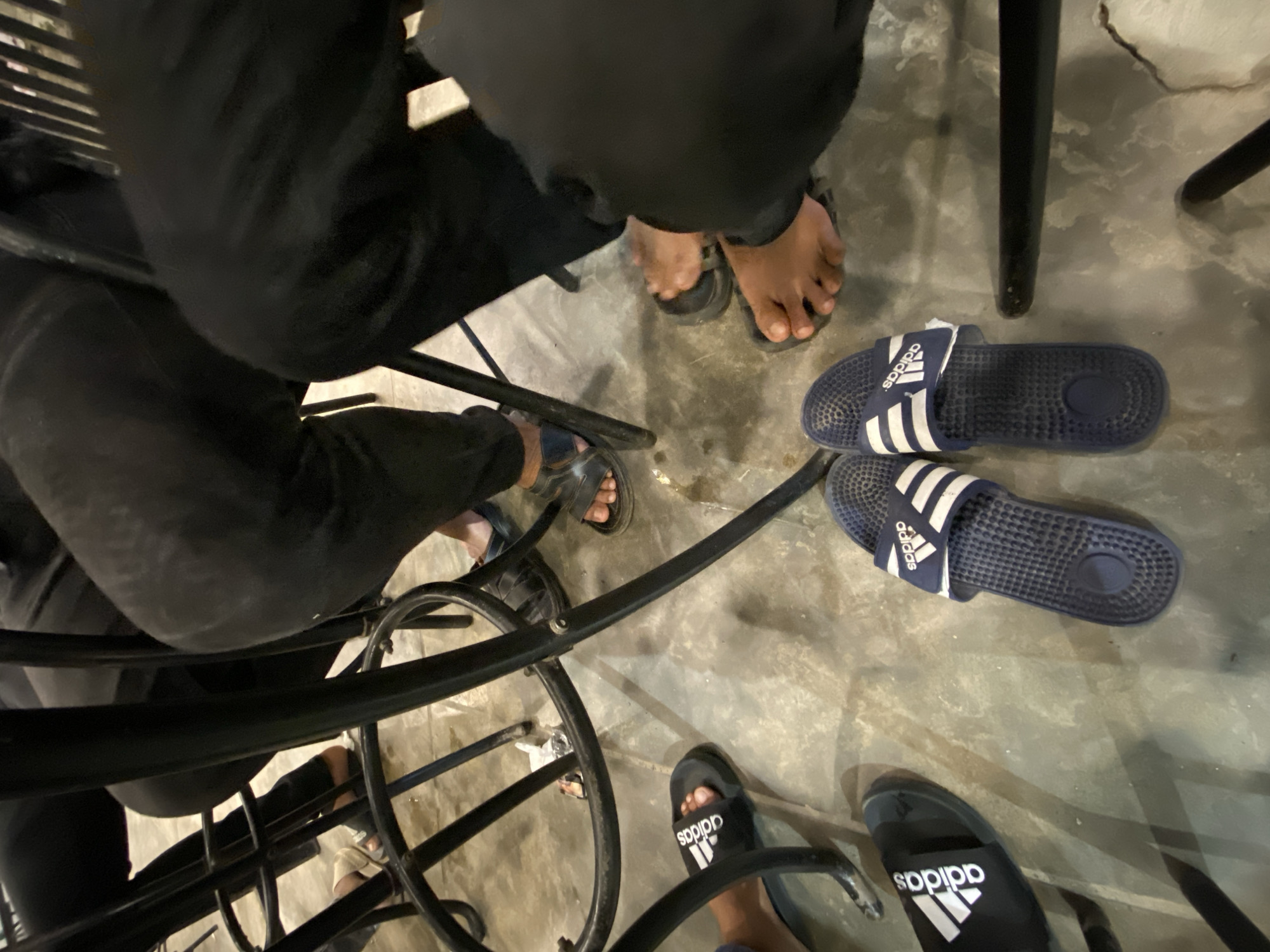 The image, taken from above, shows a group of people seated around tables in what appears to be a casual setting in Keudeublang, Indonesia. Several pairs of sandals, mostly Adidas-branded, are visible on the ground near the tables, indicating the relaxed atmosphere. The participants are wearing dark clothing, and some have removed their footwear, leaving it beneath their seats. The floor appears to be made of concrete.