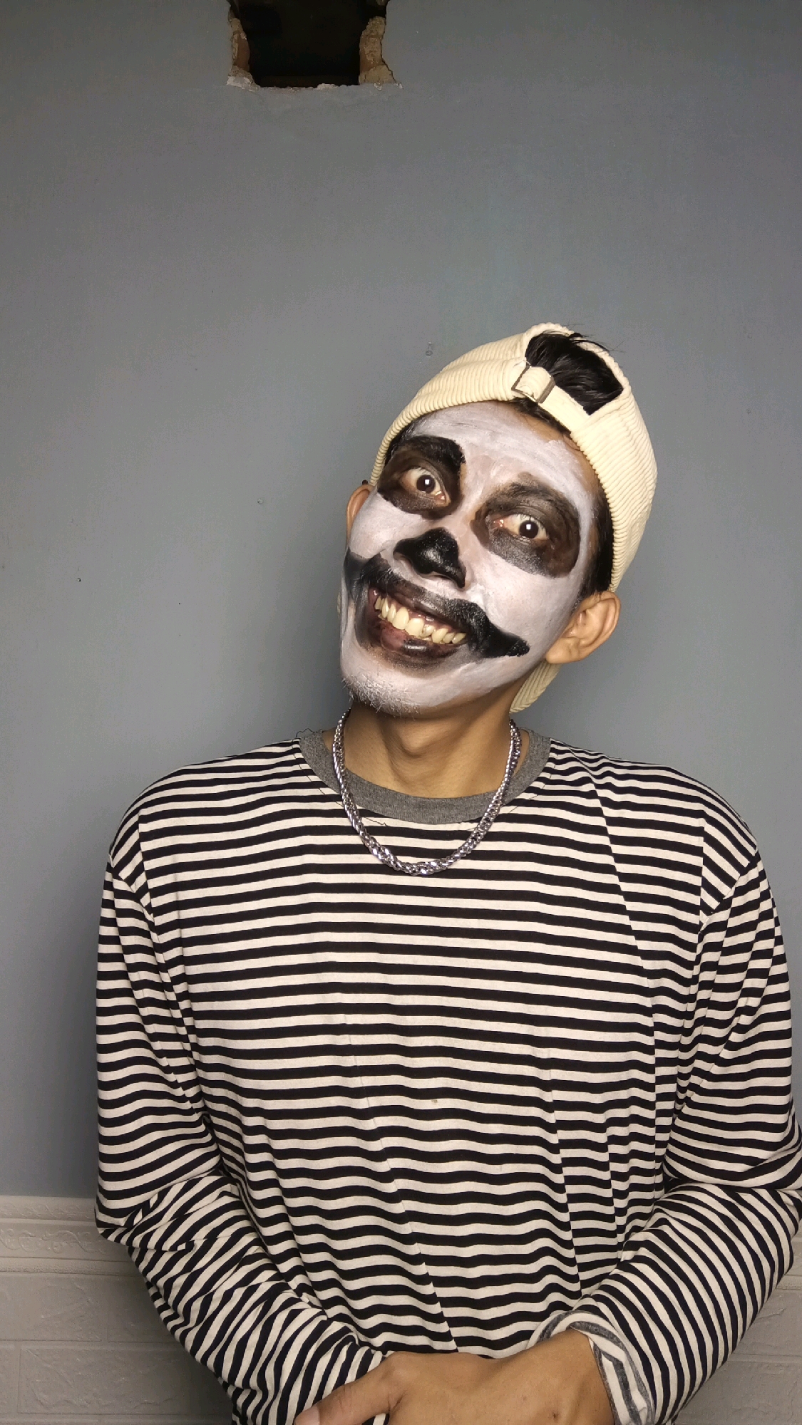 A young man with skull-like face paint and a cream-colored beanie poses against a gray wall. The paint features white on his face, black around his eyes and nose, and black lines to simulate teeth and a mustache. He is wearing a black and white striped long-sleeved shirt and a silver chain necklace. The background wall has a noticeable square hole at the top. The image appears to be taken indoors, possibly in a home or studio setting, in Kebumen, Indonesia, given the context provided.