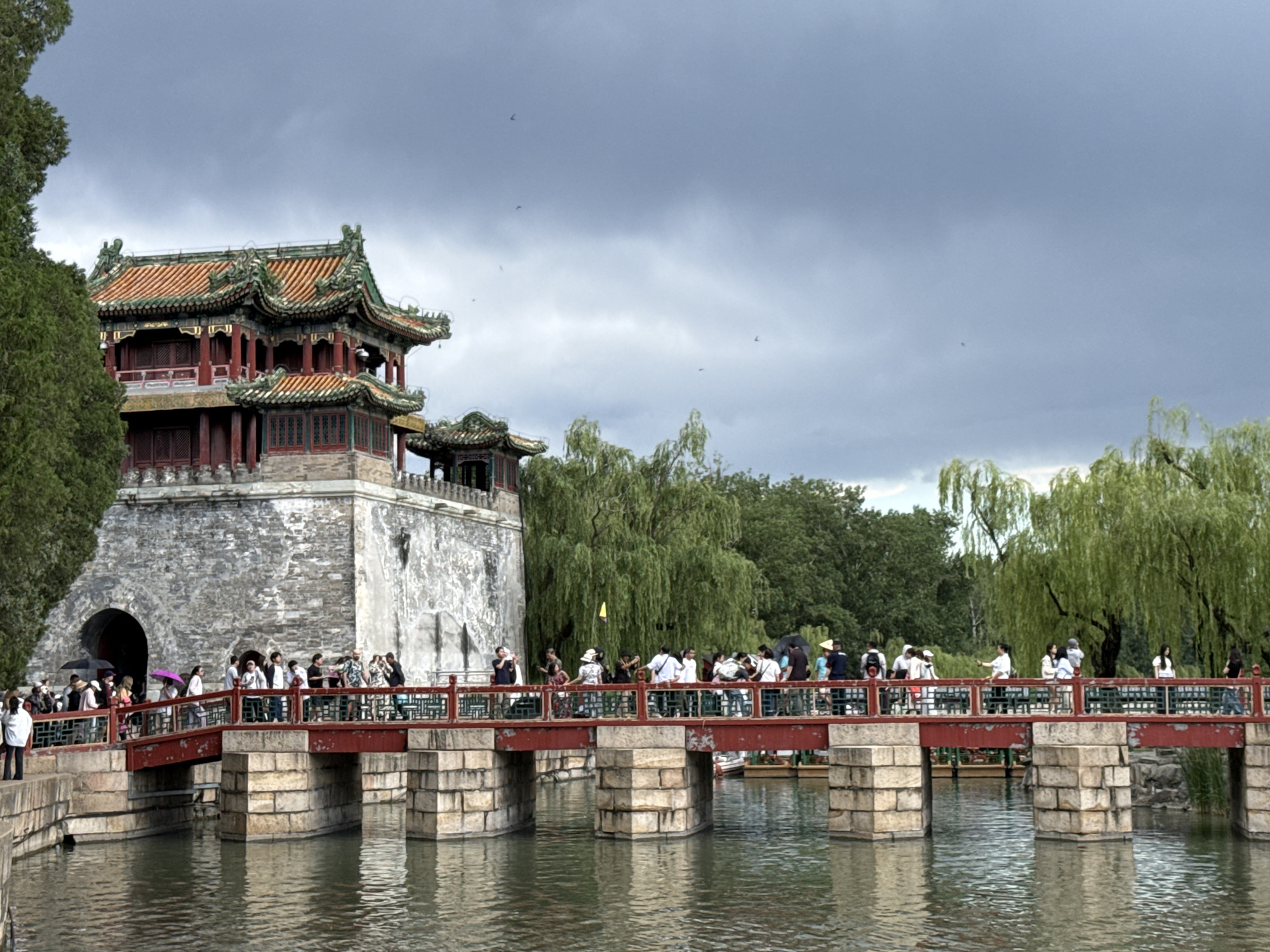 This image depicts a detailed view of the Summer Palace (Yiheyuan) in Haidian District, Beijing, China. On the left, a multi-story traditional Chinese building stands on a rough-hewn stone base, which features a dark arched entryway at its lower left. The building's upper sections consist of dark red wooden walls with multiple windows and columns, topped by elaborate, multi-tiered roofs tiled in orange-red and green, characterized by upturned eaves. A dense, dark green tree partially obscures the left side of this structure.
In the foreground, a red-painted, multi-arched stone bridge with green railings extends across a body of water, leading towards the base of the large building. The bridge is supported by multiple rectangular stone piers visible above the water line. The water surface below is calm, displaying ripples and reflections of the sky and structures.
Numerous individuals are present on the bridge, walking in both directions. They are dressed in various casual modern attire. Several people hold umbrellas, including a distinct purple one and a black one, suggesting protection from sun or light rain. In the background, beyond the bridge, lush green foliage, including several weeping willow trees, creates a verdant landscape. The sky above is overcast, dominated by dark grey clouds, and several small, dark avian silhouettes are visible against the cloud cover.