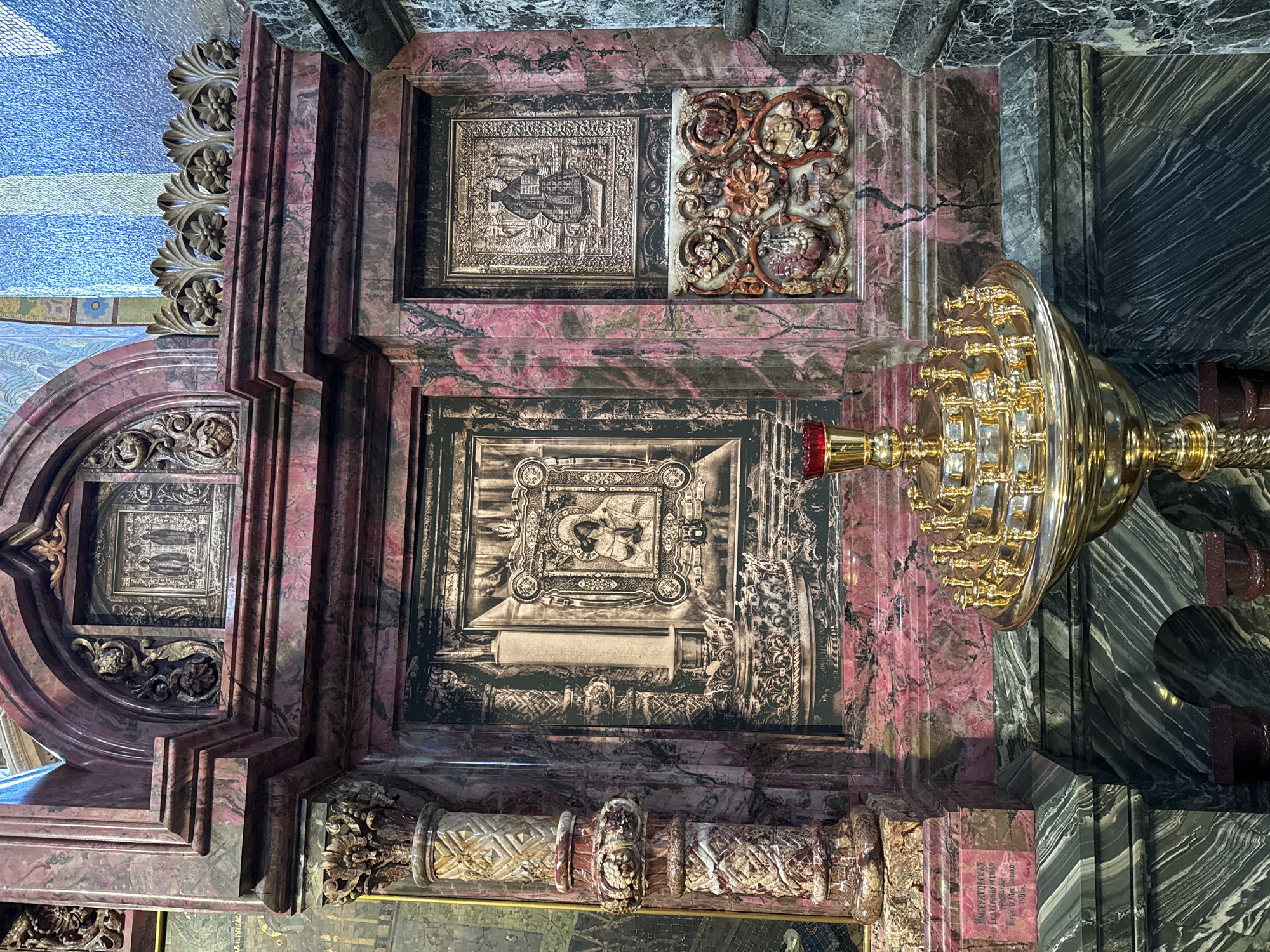 This is a close-up shot of ornate interior architectural details within a religious setting, potentially a church or cathedral. The location is Saint Petersburg, Russia.
The architectural details feature a combination of materials, primarily pink marble with dark veins, juxtaposed against dark gray/black marble. Carved embellishments in a lighter color, potentially stone or metal, adorn the structure.
The focal points are religious-themed panels or decorations. One panel presents a bas-relief carving. Another panel displays an icon or religious image.
A large golden chandelier with multiple candle holders hangs within the scene, partially obscuring some of the background details. The overall impression is of opulence, grandeur, and religious significance, reflecting the rich history and artistic heritage of Saint Petersburg.