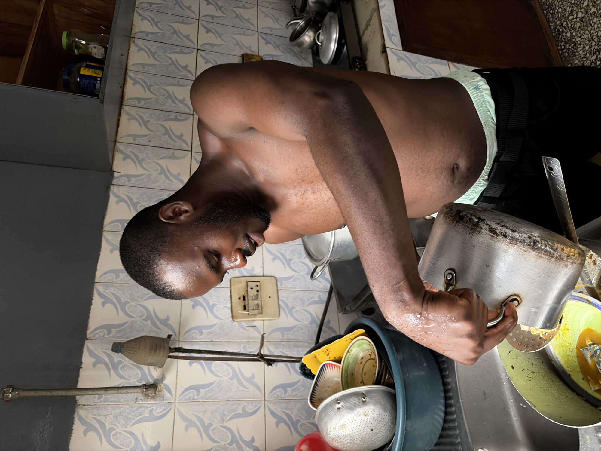 A dark-skinned man in black pants and a light blue patterned waistband is washing dishes in a kitchen. He holds a metal pot with his right hand, tilting it over a sink filled with soapy water and dirty dishes. The kitchen walls are tiled with blue and white patterned tiles. An electrical outlet is visible on the wall next to the sink. The location is Jos, Nigeria.