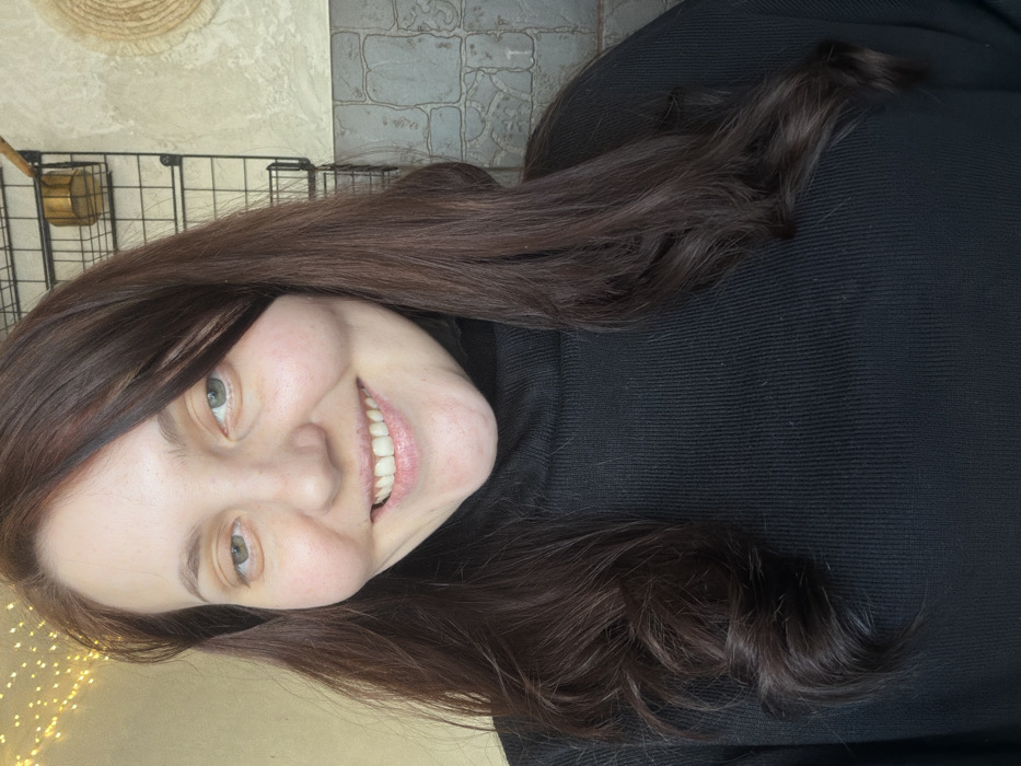 A woman with fair skin and brown hair smiles, facing the camera at an angle. Her eyes are light-colored. She wears a dark turtleneck sweater. Behind her, a wall is adorned with a woven straw mat and a black wire grid with a small gold container. A string of warm-toned lights is faintly visible in the lower left corner. The image is well-lit, focusing on the woman's face and upper body.