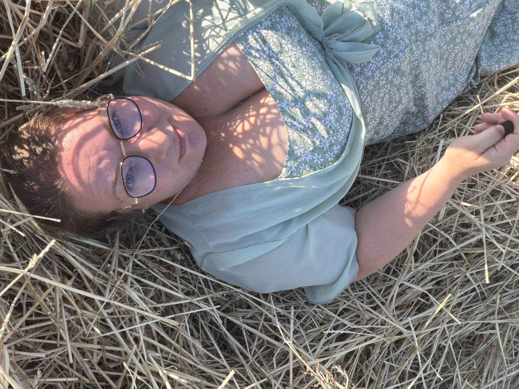 An overhead, medium shot captures a woman lying on her back in a field of dry, light-brown hay. She is positioned in the center of the frame, looking upwards towards the camera. The woman has fair skin, brown hair pulled back, and is wearing gold-rimmed glasses with blue lenses. Her attire consists of a pale green, short-sleeved dress with a ditsy floral pattern in white, and a sheer, pale green wrap or cardigan tied at the waist. A thin silver chain with a small pendant is visible around her neck. Her right arm is bent and her hand is visible near the bottom edge of the frame, holding a dark object. Sunlight casts dappled shadows across her face and chest, creating a pattern of light and dark. The background is entirely composed of the dense, rough texture of hay. The location is Ufhusen, Switzerland.