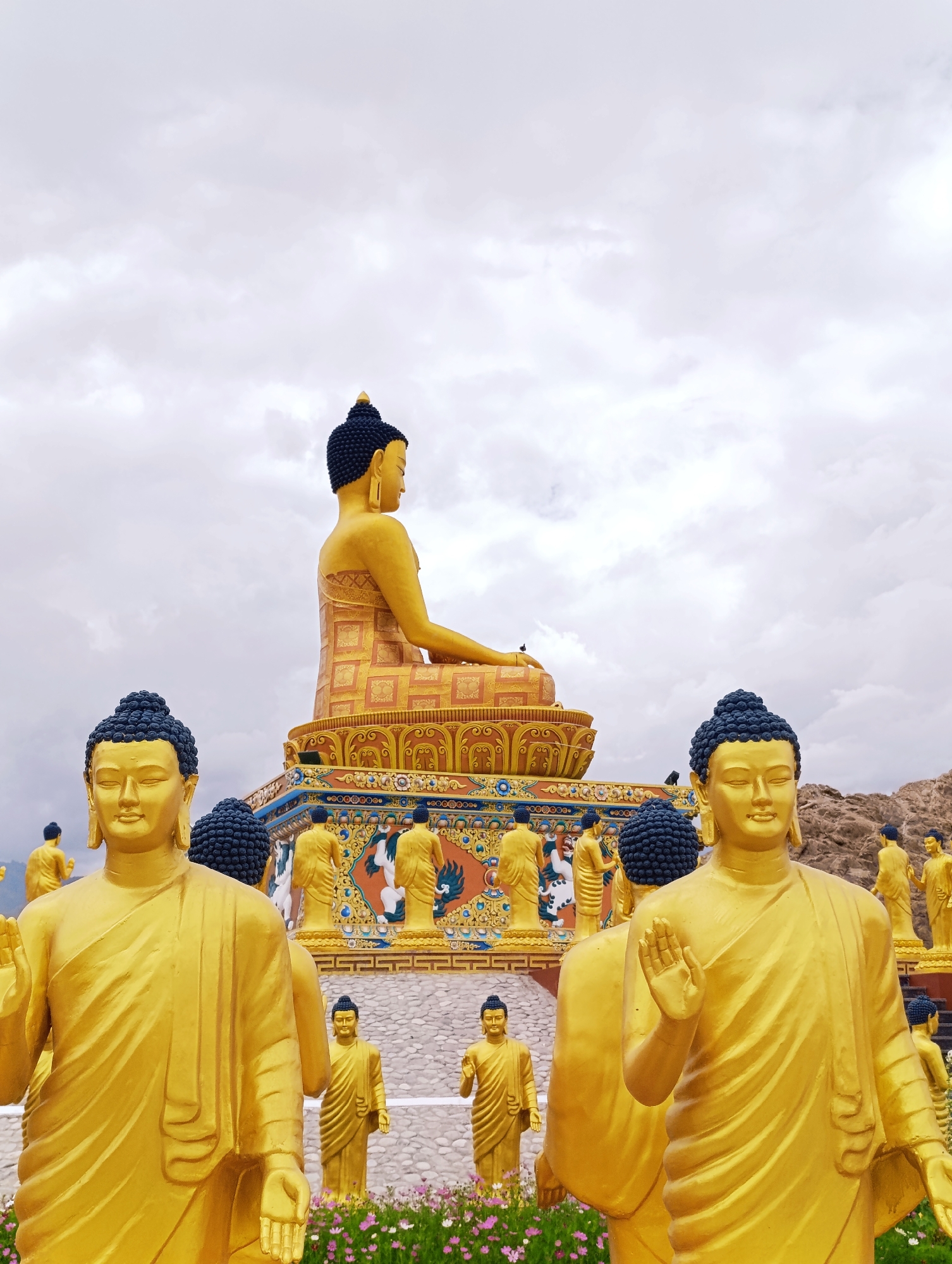 In Leh, India, a monumental golden Buddha statue sits atop a multi-tiered platform adorned with intricate carvings and smaller figures. In the foreground, several life-sized golden Buddha statues stand, facing forward with serene expressions and raised right hands in a gesture of blessing. These statues are surrounded by a low wall of grey stones and a vibrant bed of pink and purple flowers. The sky above is overcast with a soft, diffused light. The scene appears to be part of a Buddhist monastery or temple complex.