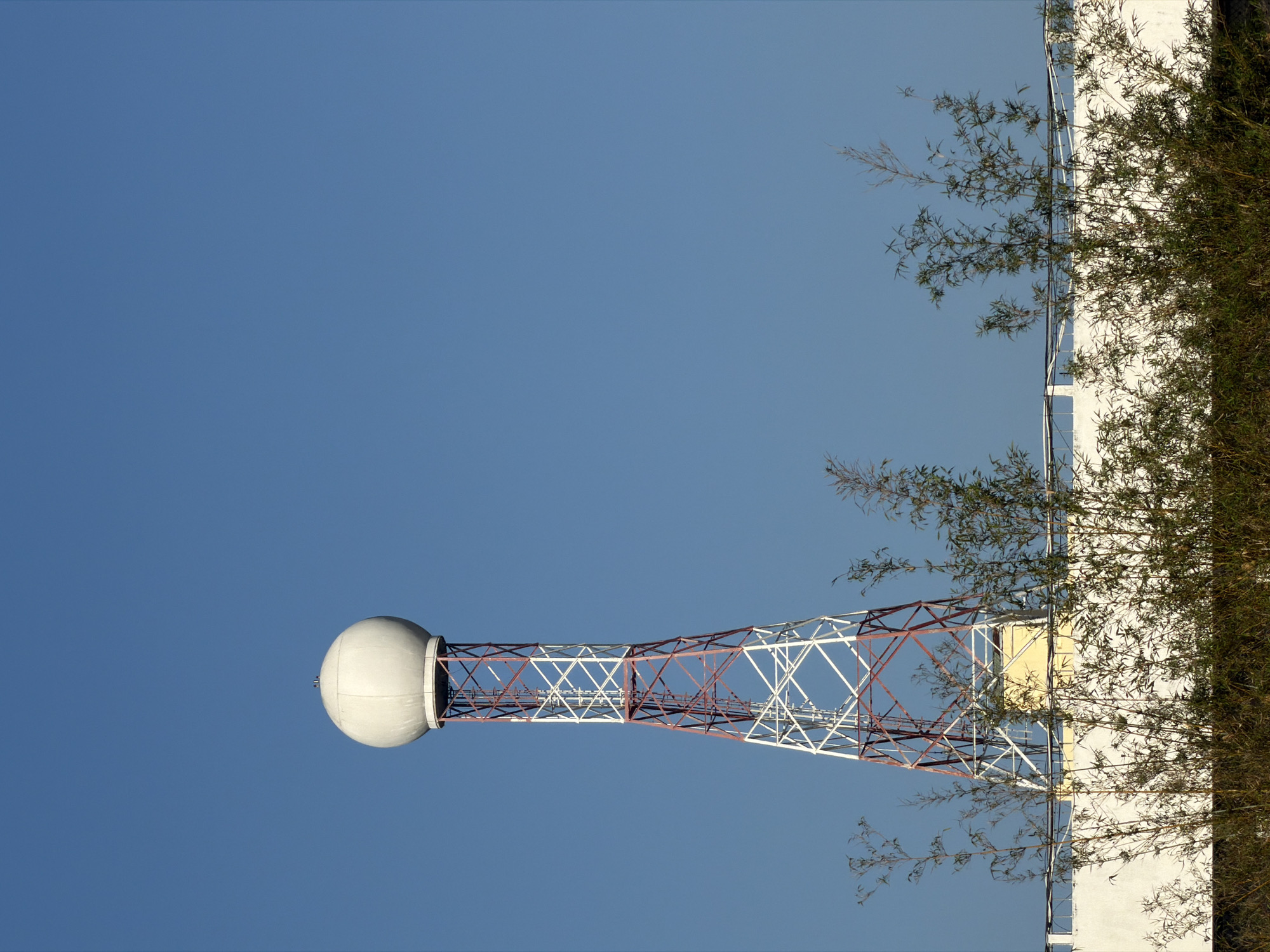 The image depicts a weather radar tower located in Nha Trang Ward, Vietnam, under a clear, cloudless blue sky. The tower has a large, white spherical radome at its top, supported by a lattice structure painted with alternating red and white sections. The base of the tower is connected to a white building. A dense patch of bamboo-like vegetation partially obscures the view of the building and extends upwards along its side.