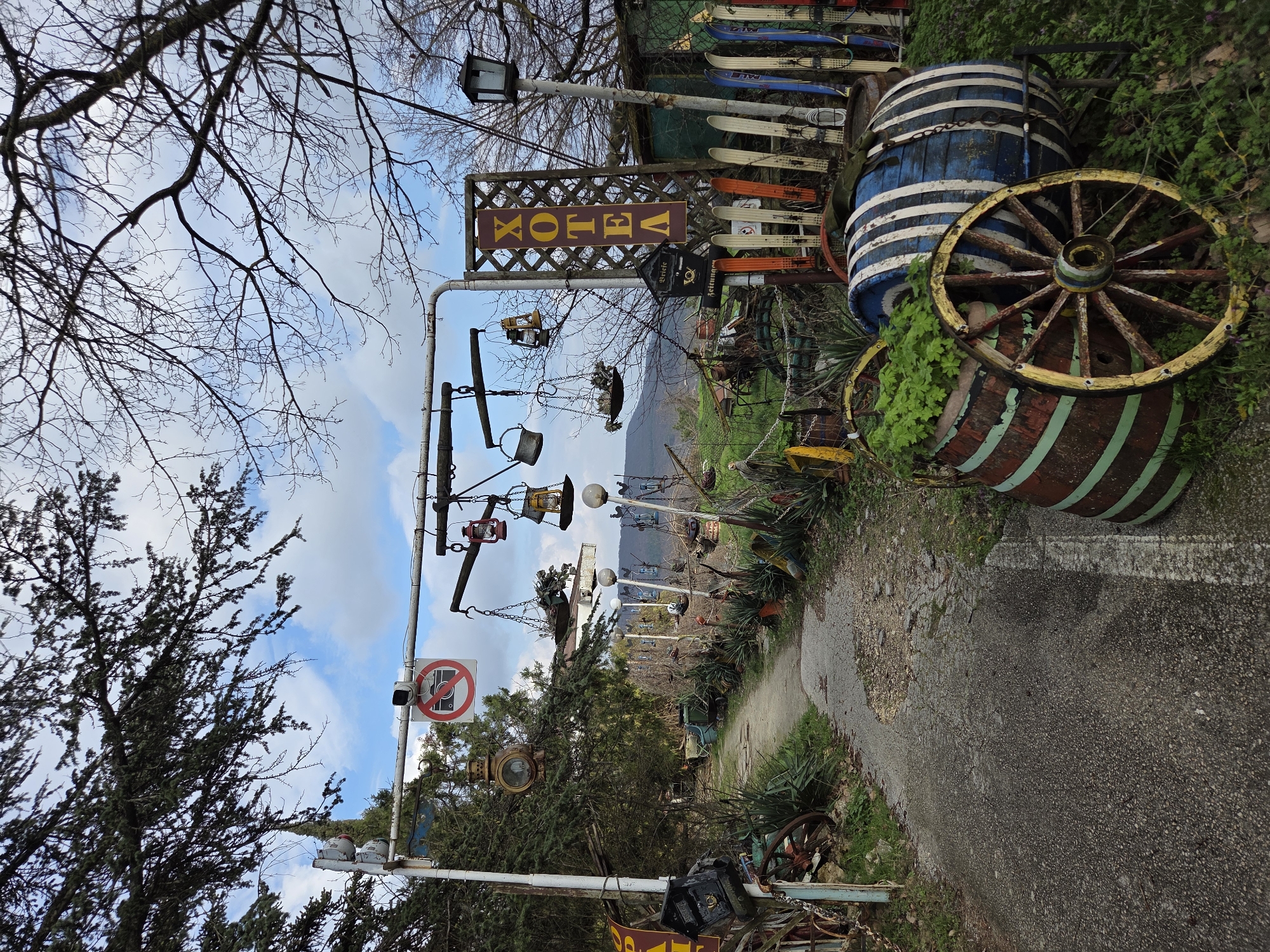 The image depicts an outdoor area, possibly a garden or yard, in the city of Aksakovo, Bulgaria. The scene is characterized by a mix of decorative and seemingly discarded objects. There are several barrels, some painted with stripes and others used as planters, as well as wooden wagon wheels. Skis are stacked up in the background. A metal structure holds various lanterns and decorative items, including a sign with lettering in what appears to be Cyrillic script. A "no photography" sign is visible on the metal structure. The ground is a mix of pavement and dirt, with vegetation growing along the edges. Bare trees are seen in the background, suggesting it is not high summer. The sky is partly cloudy.
