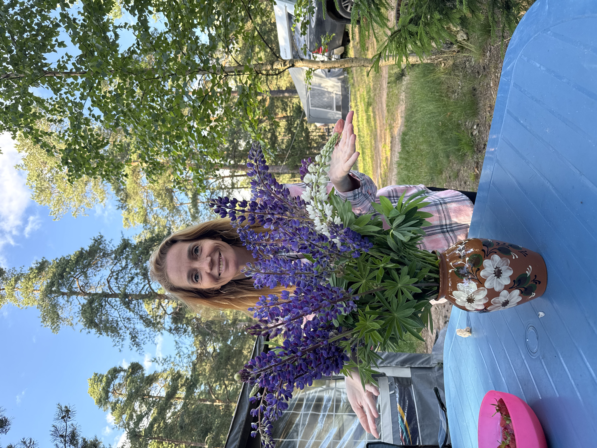 A woman with shoulder-length blonde hair and a light pink and white plaid shirt stands in a forest clearing, smiling at the camera with her hands outstretched, palms up. In front of her, on a bright blue plastic table, is a brown ceramic vase filled with a bouquet of purple and white lupine flowers. To the left of the vase is a bright pink bowl containing a pile of red strawberries. In the background, a camping trailer and a dark SUV are visible, along with tall pine and birch trees. The scene appears to be set in Zelenaya Roshcha, Russia, under a bright blue sky with scattered clouds.