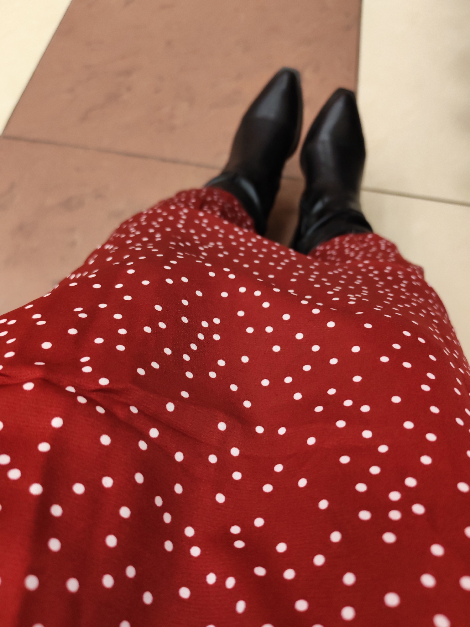 The image provides a first-person, downward-facing perspective of a person's legs and feet, likely taken from a seated or standing position looking towards the ground. The main subject is a vibrant red dress or skirt covered in numerous small, irregularly spaced white polka dots. The fabric appears to be soft and flowing, gathered around the upper legs.
The person is wearing a pair of sleek, black, pointed-toe ankle boots, which cover their feet and lower calves. The footwear and garment suggest a fashionable and contemporary outfit.
The immediate environment consists of a clean, tiled floor with two distinct sections. To the upper left, the floor is a solid, warm reddish-brown or terracotta color. To the right and lower section, the floor is a lighter, creamy beige or off-white. The clear division between these two floor colors suggests an indoor public or commercial setting, such as a shopping center, office building, or an airport, in Izhevsk, Russia.
No other people, objects, or text are visible in the frame. The lighting is consistent with indoor ambient light. The image captures a personal moment, emphasizing the attire and the immediate surroundings from the wearer's point of view, without revealing specific external details or emotions.