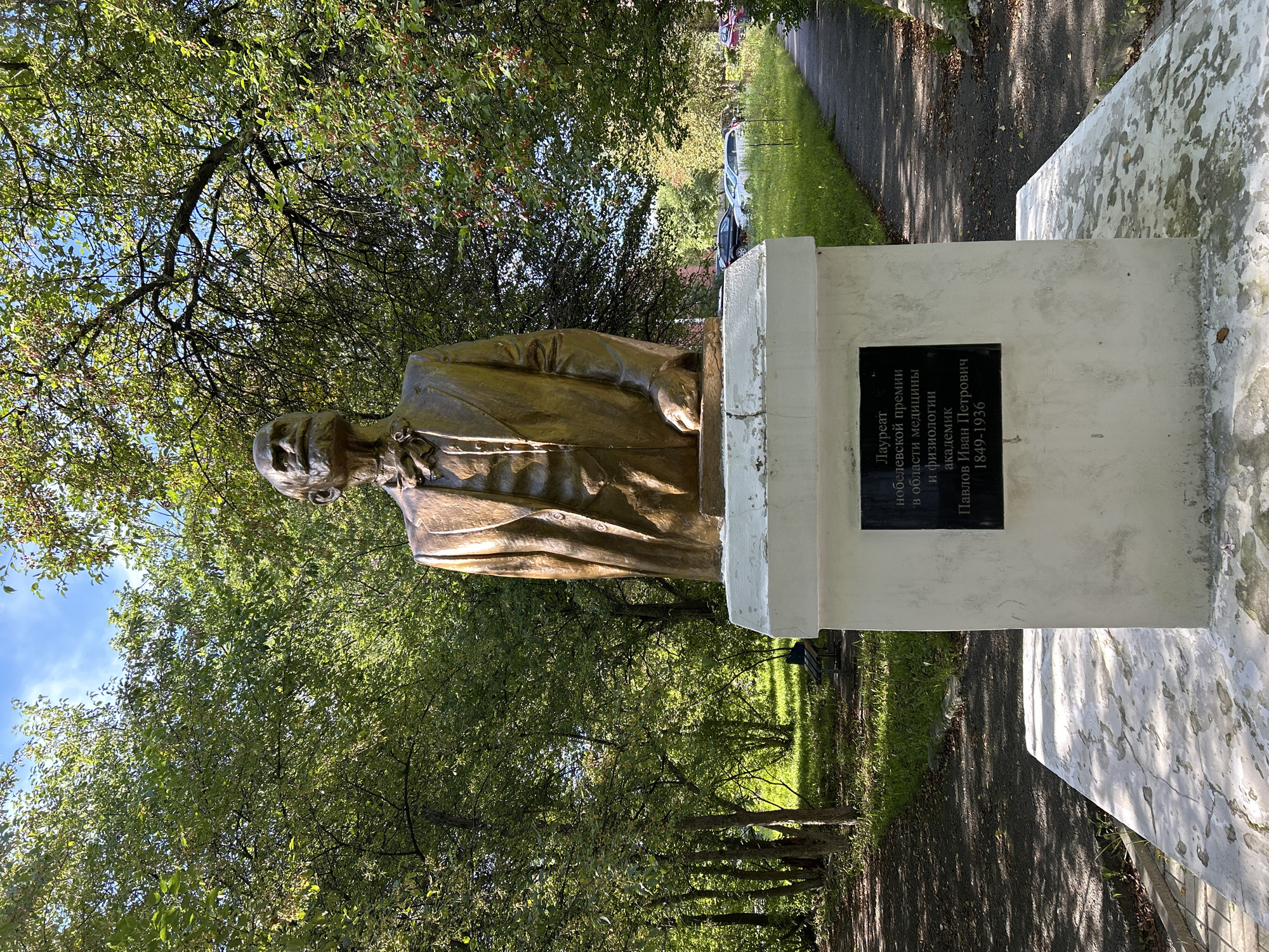 The image shows a bronze bust of a man with a mustache, wearing a suit and tie, on a white pedestal. A black plaque is attached to the pedestal with Russian text. The bust is located outdoors, with trees and foliage in the background. A paved walkway and a grassy area with parked cars are also visible. The plaque reads "Laureate of the Nobel Prize in Physiology or Medicine, Academician Pavlov Ivan Petrovich, 1849-1936." This indicates the monument is dedicated to Ivan Pavlov, a Russian physiologist. The scene is consistent with the monument located on the territory of Siberian State Medical University in Tomsk, Russia, where Pavlov was a student and later a professor.